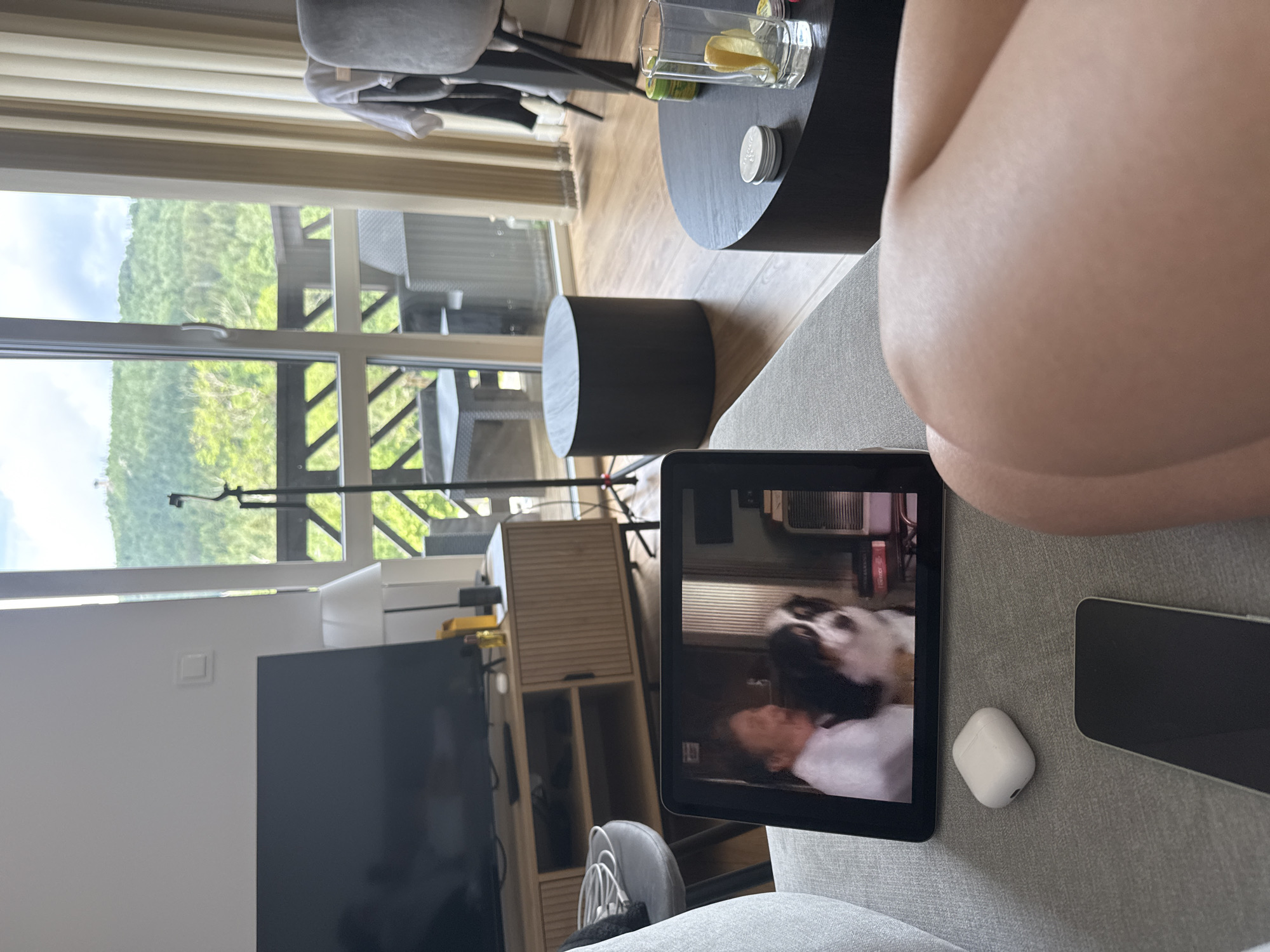 In a brightly lit room, a person is seated on a sofa, with their legs propped up on a coffee table. The room is oriented towards a large window that offers a scenic view of lush green mountains, suggesting a location in Szklarska Poręba, Poland. A television is visible on the left, and a tablet in front of the person displays a video of a person interacting with a dog, possibly a Bernese Mountain Dog. Nearby, a smartphone and a pair of wireless earbuds rest on the sofa. The atmosphere appears relaxed and casual, hinting at a leisurely afternoon. The weather outside seems pleasant, with visible sunlight and a clear sky. The room itself is modern, with minimalist furniture and a light-colored wooden floor.