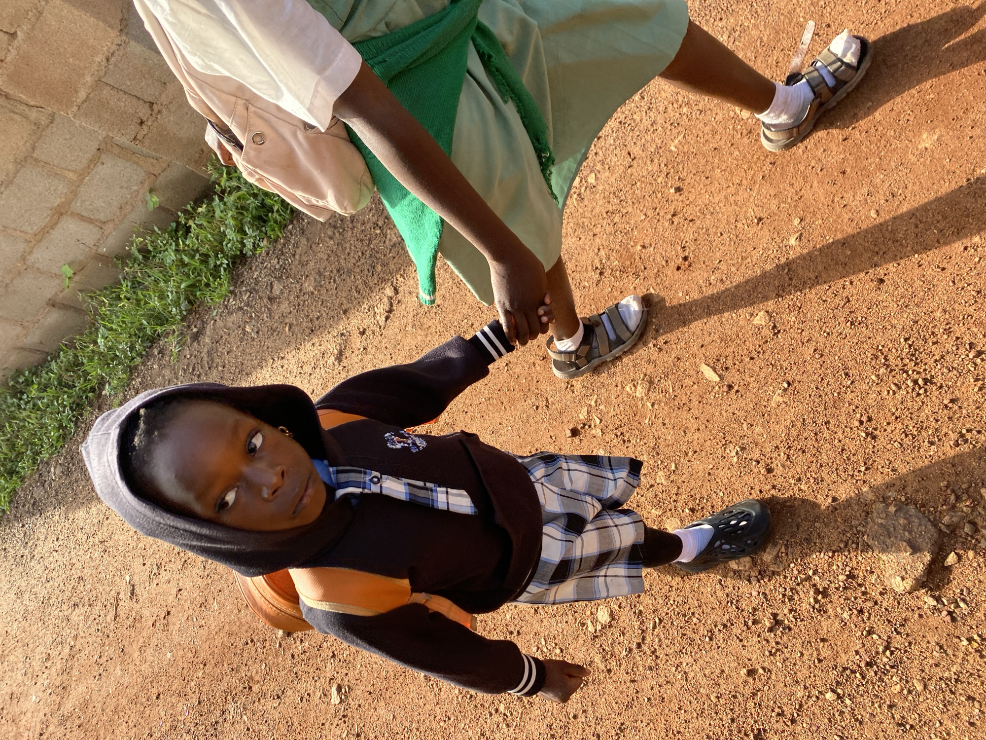 In the city of Bukuru, Nigeria, a young child wearing a hooded black jacket with an orange backpack, plaid shorts, and black perforated shoes stands on a dirt path. The child is looking to the left, with a slight frown. Another person, wearing a light-colored shirt and a light green skirt, is reaching out to the child, holding the child's hand. The background consists of a brick wall and green foliage. The scene is illuminated by sunlight, casting shadows on the ground.