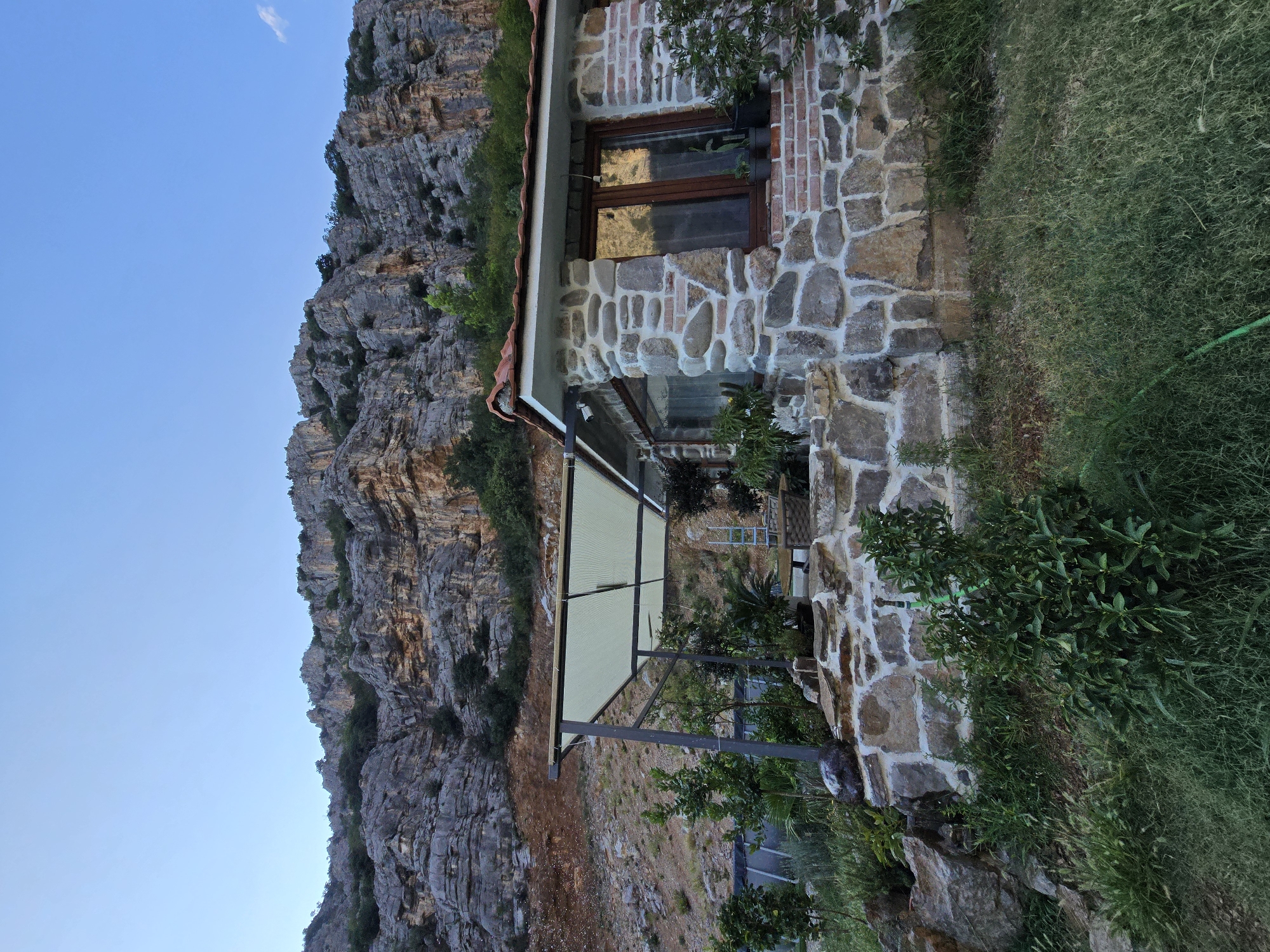 A low-angle, full shot captures a rustic stone cottage nestled against a towering, rugged cliff face under a clear blue sky. The cottage, constructed from irregular, grey and tan stones with visible white mortar, features a red-tiled roof and a large wooden-framed window. To the left of the cottage, a light-colored canvas awning is supported by dark metal poles, covering a seating area with wicker furniture and potted plants. The surrounding landscape is a mix of green grass, small trees, bushes, and some larger rocks. The cliff face behind the cottage is steep and rocky, with patches of vegetation clinging to its surface. The overall impression is one of a remote, secluded dwelling in a natural, mountainous environment, likely in the outskirts of Podgorica, Montenegro, given the characteristic karst landscape. There are no people present in the image.