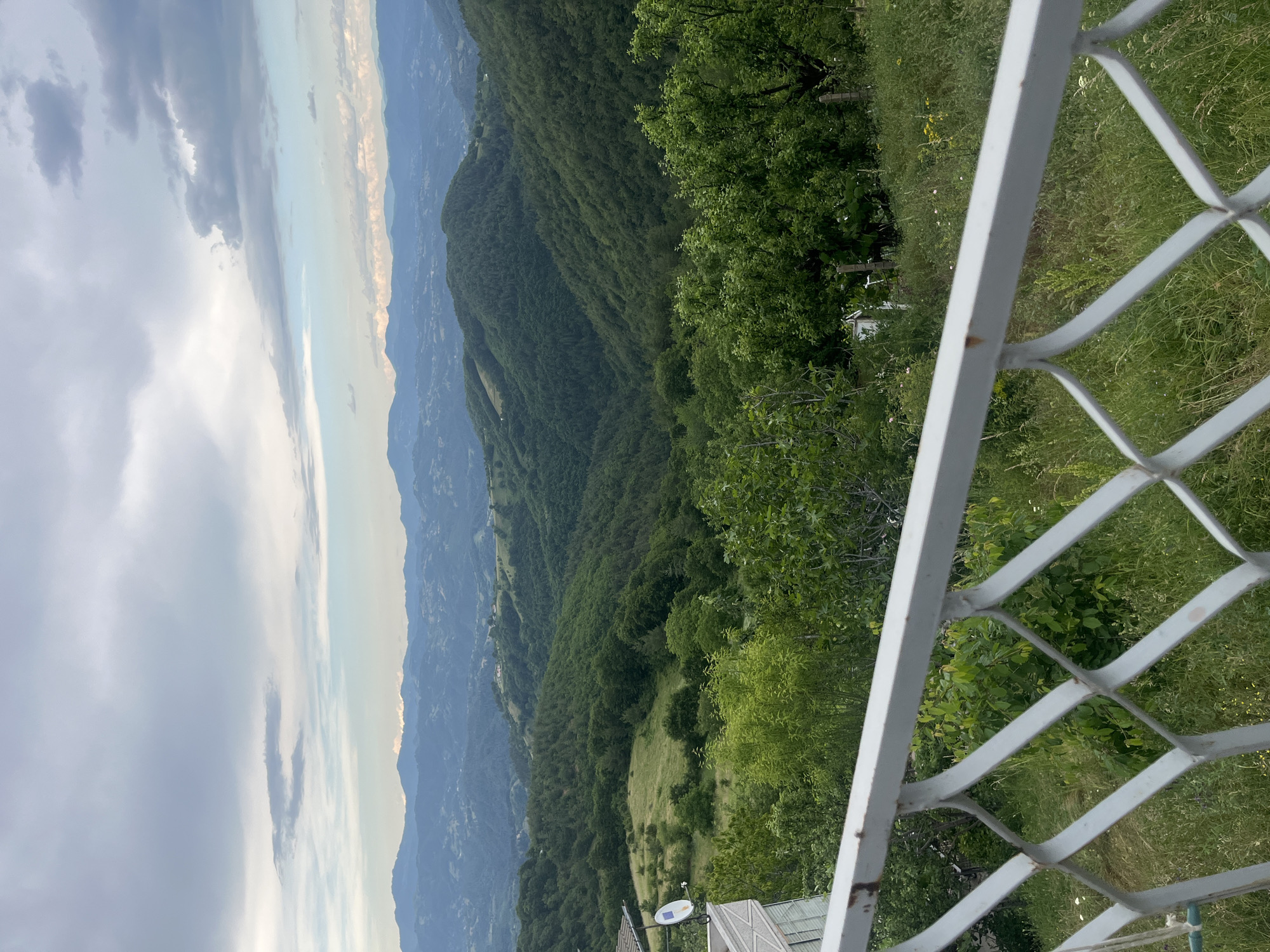 The image displays a panoramic view of rolling green hills and mountains under a cloudy sky. In the foreground, a white metal railing with a diamond pattern traverses the bottom of the frame, obscuring some of the vegetation and a portion of a building with a satellite dish. The distant landscape is characterized by layers of forested hills and mountains fading into a hazy horizon. The scene appears to be a scenic overlook in the rural area of Startsevo, Bulgaria.