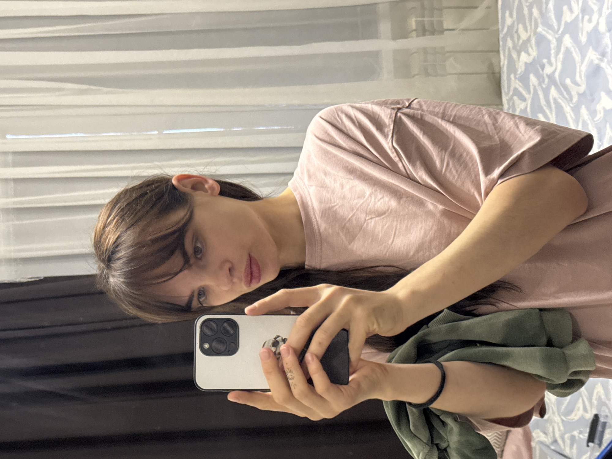 A woman with brown hair and bangs is holding a white iPhone in front of her face. She is wearing a pink t-shirt and holding a green garment, possibly a hoodie or jacket, with her right hand. The background consists of a window with white blinds and a patterned blue and white bedspread. The setting is likely indoors, but the location in Rostov-on-Don, Russia, cannot be determined from the image.