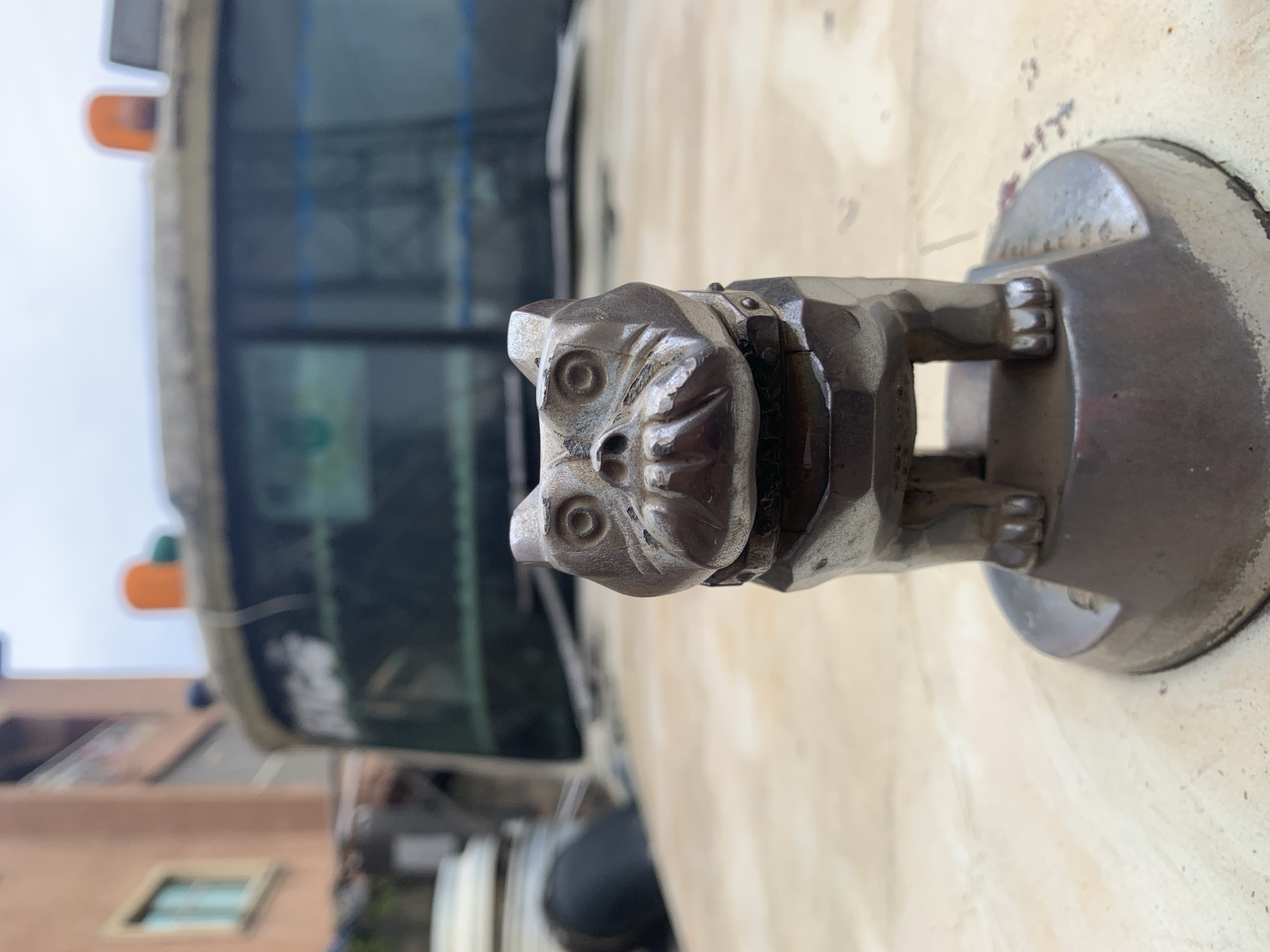 The image shows a close-up of a metal bulldog hood ornament affixed to the hood of a vehicle. The ornament is weathered and appears to be made of a dull silver-colored metal. It depicts a bulldog with a wrinkled face, bulging eyes, and an open mouth showing teeth. The ornament is mounted on a circular base. In the background, out of focus, is the windshield of the vehicle, with some writing on it. Above the windshield, two amber hazard lights are visible. Further in the background, a building with an orange facade and a window is discernible. The overall environment suggests an outdoor setting in a city, possibly in Nkpor, Nigeria, given the context of the location. The hood of the vehicle is a light beige color and shows some signs of wear.