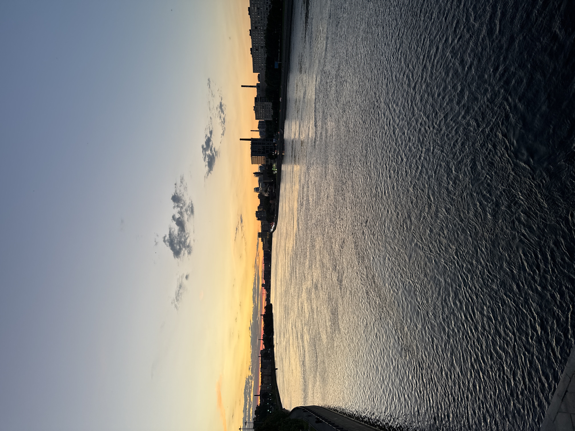 This is a horizontal, eye-level photograph capturing a wide river at sunset. The sky transitions from a pale blue at the top to a vibrant orange and yellow near the horizon, with a few scattered clouds. On the left bank of the river, a curved embankment with a walkway and trees is visible, leading towards buildings in the distance. The right bank features a cityscape with various buildings, including some with smokestacks, and streetlights. The river's surface is dark and reflects the colors of the sky, with visible ripples and waves. The overall impression is a peaceful urban scene at dusk. While the prompt mentions the city of Romanovka, Russia, there are no specific visual cues in the image that definitively confirm this location.