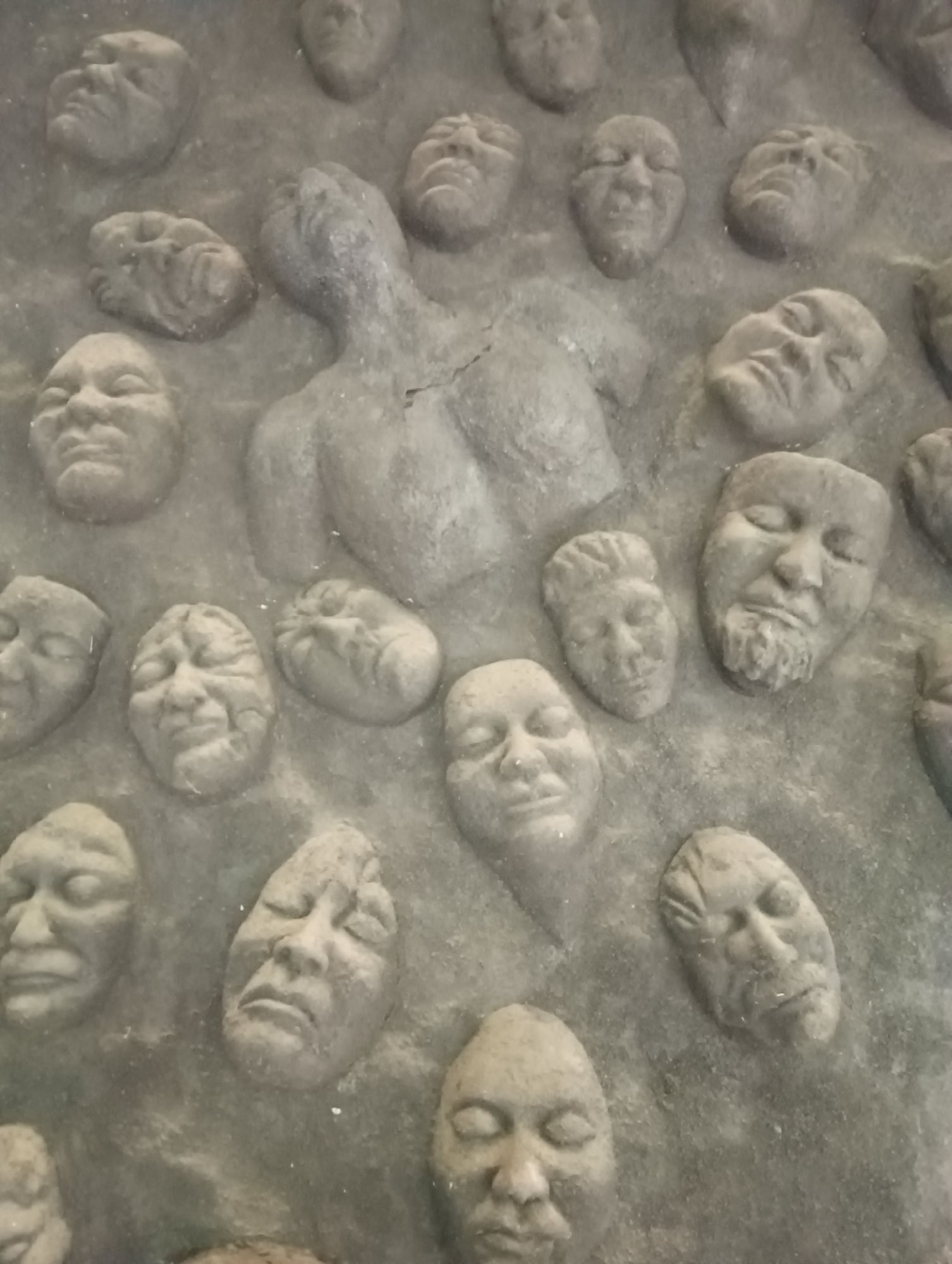 This image depicts a section of a sculpture located in Old Manali, India, featuring a collection of carved faces embedded in a textured surface. The sculpture appears to be made of concrete or stone, with a rough, weathered finish. The faces are varied in their expressions and apparent age, with some appearing as children, others as adults, and one central, larger figure that seems to be a torso with a head tilted back. The individual faces are rendered with distinct features, including eyes, noses, and mouths, though some details are softened by the texture of the material and the lighting. The overall impression is one of a crowd or a collection of souls, perhaps representing a community or a historical narrative. The background is uniform, suggesting it's a wall or a large panel. The lighting creates shadows that highlight the contours of the faces and the texture of the sculpture.