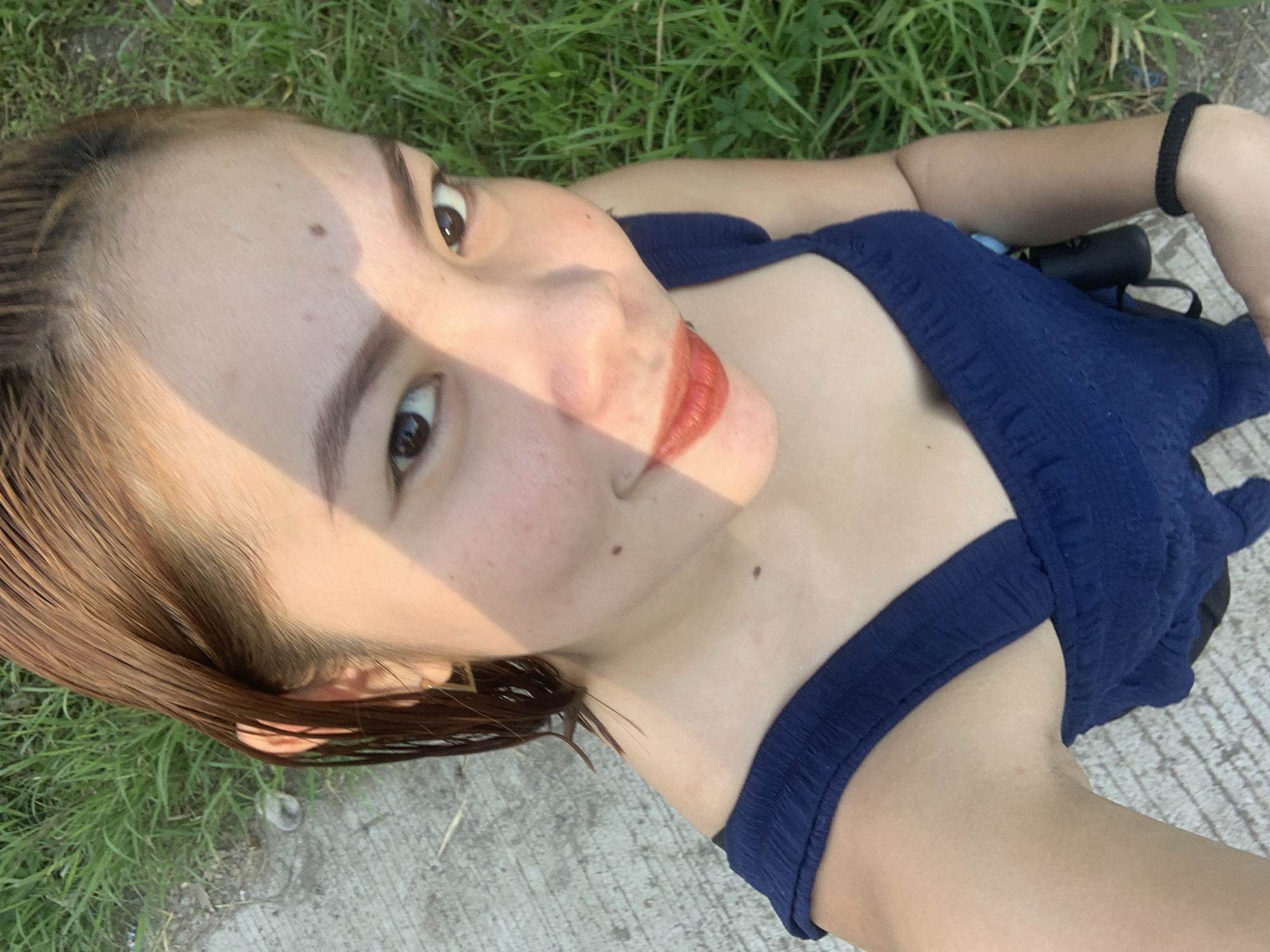 The media file features a close-up, top-down shot of a young woman lying on her back outdoors. She has light brown hair, styled to fall around her face, and her head is tilted to her left. Her eyes are open and looking towards the camera, and she is wearing coral-colored lipstick. A shadow, possibly from sunlight filtering through leaves, casts a diagonal line across her face from her forehead down to her cheek. Her skin appears fair with a scattering of freckles on her cheeks and a mole on her right cheek. She is wearing a dark blue, sleeveless top with textured fabric. Her right arm is bent, with her hand resting near her head, and she wears a black elastic band on her wrist. Her left arm is also bent, and the image crops to show her upper chest and shoulder. The background consists of green grass and a concrete surface, suggesting an outdoor setting. The lighting indicates it is likely daytime, possibly late afternoon given the warm tones of the shadow. The overall mood appears relaxed and casual, possibly a selfie taken while enjoying a quiet moment outdoors in Kiblawan, Philippines.