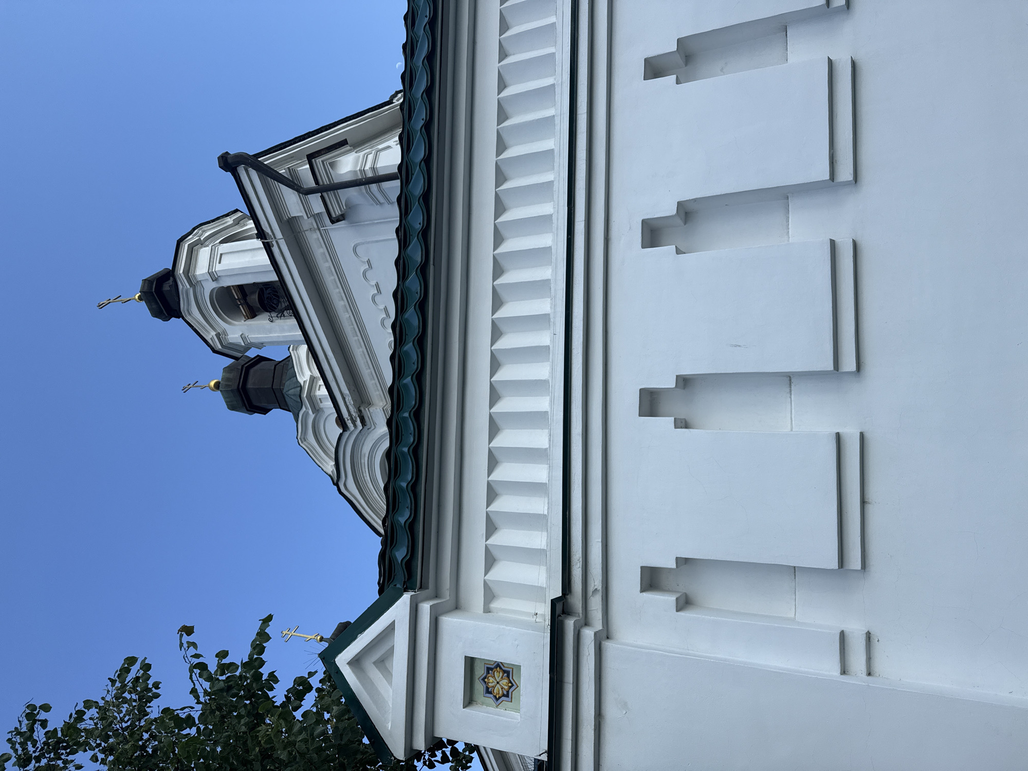 The image shows a low-angle view of the white wall and roof of a building, likely a church, in Yekaterinburg, Russia. The wall features decorative relief patterns and a small tiled mosaic. The roof is green and curved, with architectural elements and a golden cross visible against a clear blue sky. A tree branch with green leaves is visible in the upper left corner.
