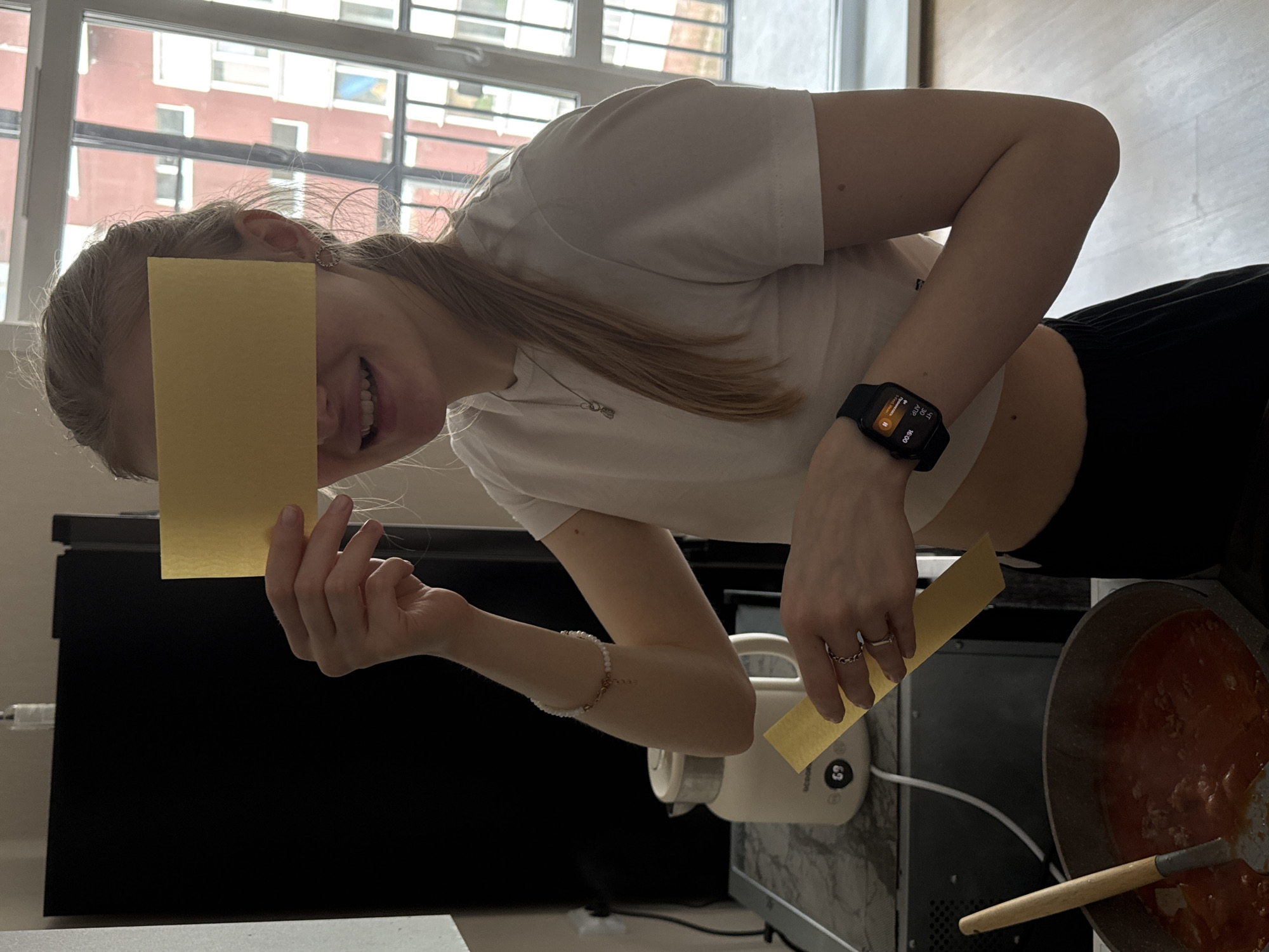 A young woman is captured in a vertical, brightly lit indoor shot, appearing to be in the process of preparing a meal, likely lasagna. Her face is partially covered by a golden-yellow lasagna pasta sheet held in her left hand, but a wide smile is visible. She holds another pasta sheet in her right hand, positioned above a pot of red meat sauce, which is being stirred with a wooden spoon.
She is dressed in a white t-shirt and dark pants, with long, light-colored hair falling over her shoulder. Her accessories include a small, circular earring, a delicate silver necklace, a white beaded bracelet on her left wrist, and two rings on her right hand. A black smartwatch on her left wrist displays the Spotify logo, text "ALAY", "AMP", and the time "16:00".
The background features a large window with a view of multi-story buildings, suggesting an urban setting. Bright daylight illuminates the scene, indicating daytime. A white appliance, possibly a kettle or rice cooker, with the brand name "DECHICO" and the number "6" visible, sits on a counter next to the pot. The overall mood is cheerful, with the woman's smile suggesting enjoyment of the activity. The location is Perm, Russia.