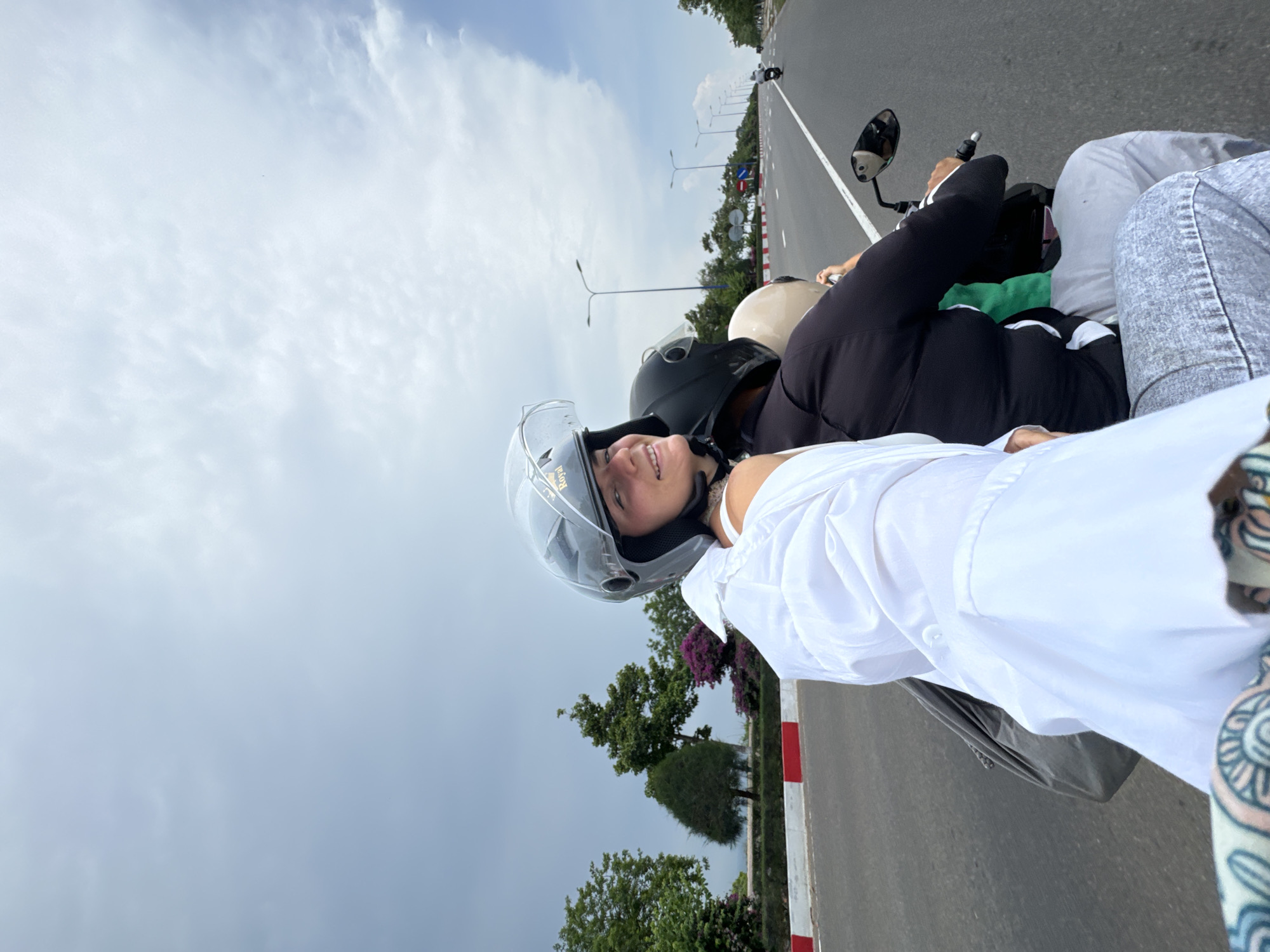 In Mũi Né, Vietnam, two individuals are riding on the back of a motorcycle. The person in front, wearing a black helmet, is operating the vehicle. Behind them, a woman with a gray helmet is seated, smiling and looking towards the camera. She is wearing a white shirt and has a backpack slung over her shoulder. A third person, wearing a beige helmet, is also seated on the motorcycle, between the driver and the woman with the gray helmet. They are all on a paved road with a red and white painted curb on the left side, suggesting a coastal or seaside location. Trees and foliage line the side of the road, and a bright, cloudy sky is visible above. In the distance, other motorcycles can be seen on the road.