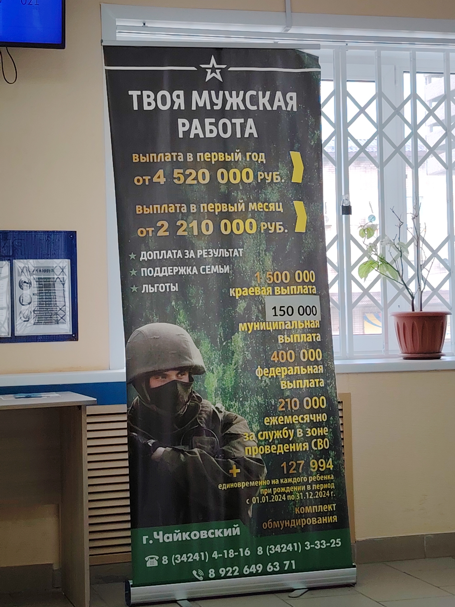 An indoor scene in Chaikovsky, Russia, features a vertical recruitment banner positioned prominently to the right of the frame's center. The banner, predominantly dark grey with a green camouflage background image, advertises "ТВОЯ МУЖСКАЯ РАБОТА" (YOUR MAN'S WORK) and details various financial incentives and benefits in Russian Rubles (RUB) for military service, including a first-year payment from 4,520,000 RUB and a first-month payment from 2,210,000 RUB, along with regional, municipal, federal, and monthly payments for service in the SVO zone, and a one-time child birth payment for 2024. An image of a soldier in a helmet and face mask is depicted at the bottom of the banner. The banner lists "г. Чайковский" (Chaikovsky city) and three phone numbers at its base.
To the left of the banner, a light beige wall with a blue horizontal stripe at the bottom features a blue-framed bulletin board displaying several documents under clear covers. Above this, a small blue digital screen shows "021". A light-colored wooden table or counter is partially visible in the lower-left foreground. To the right of the banner, a white-framed window with white accordion-style security bars is visible. A potted plant with sparse green and white leaves and bare branches sits on the windowsill in a terracotta-colored pot. The exterior view through the window is blurred, showing distant structures and a pale sky. The floor is dark grey or brown.
