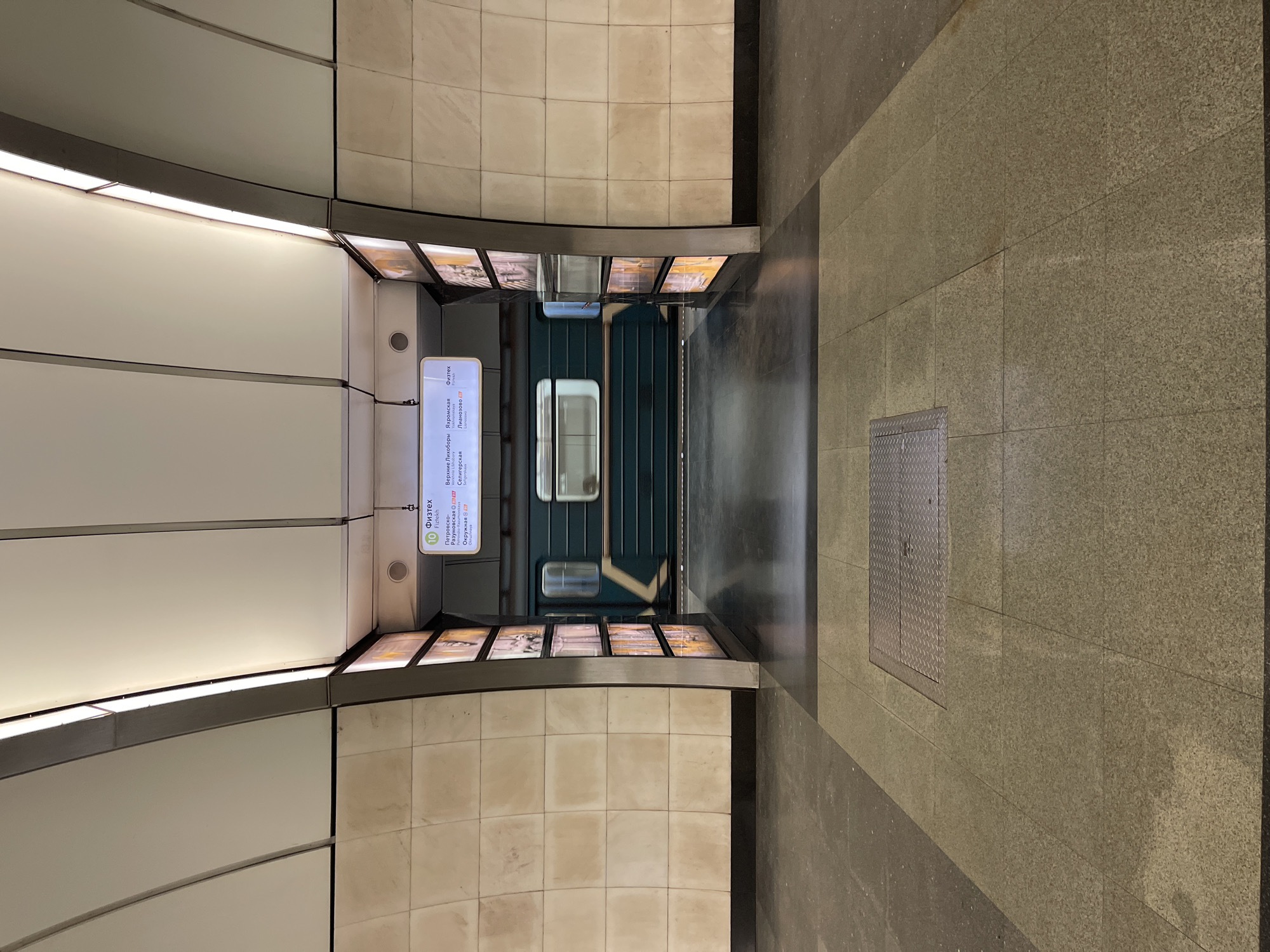 The image shows a metro station platform. A train car with dark green exterior panels and white-framed windows is centered at the back of the image. A station sign showing metro line information is mounted on the left wall, while a series of framed pictures runs along the top of the tiled walls. The floor is made of stone-like tiles with a shiny strip running down the center. A metal grate is in the floor. The location is a metro station, city of Unknown, Unknown.