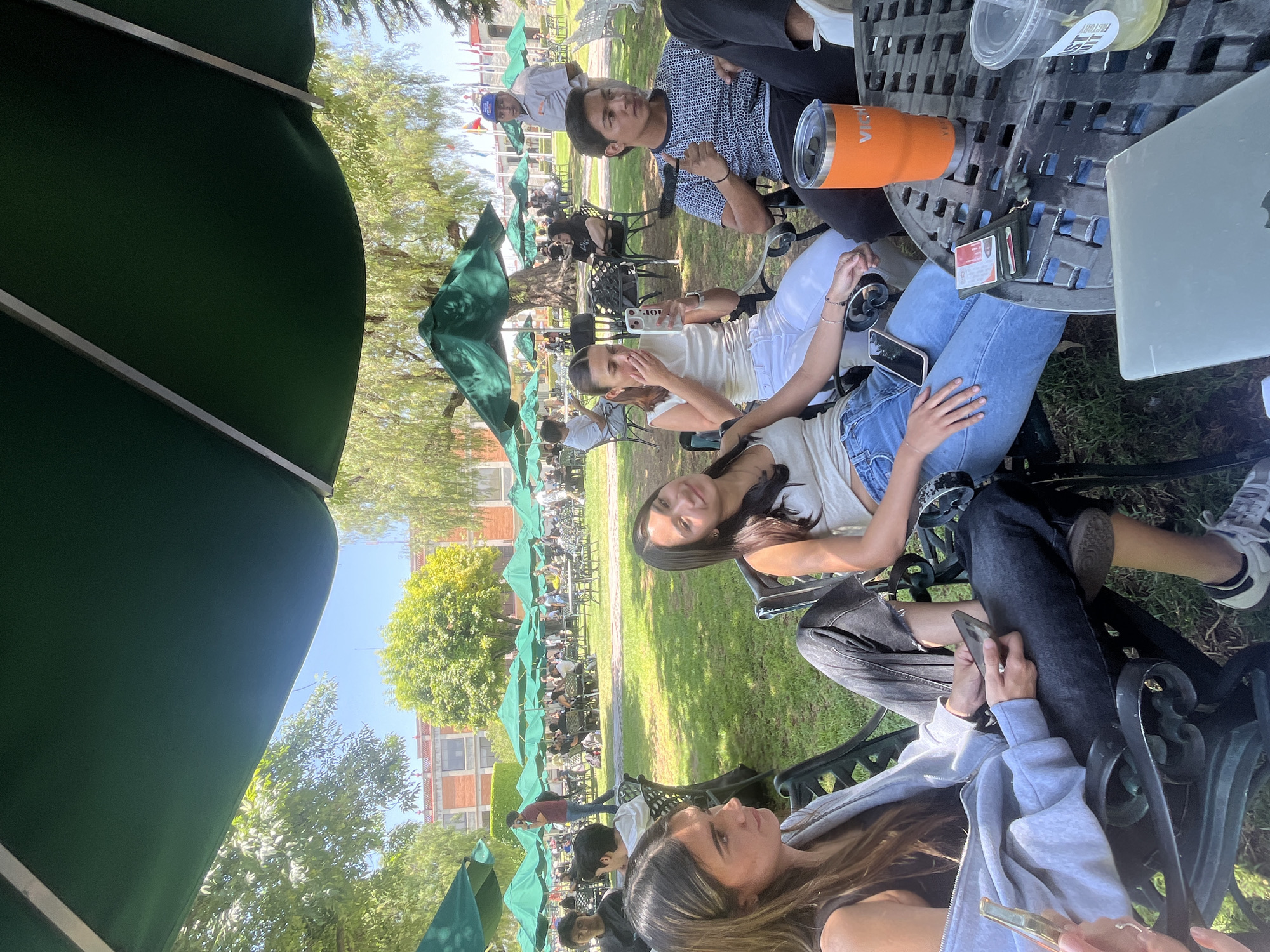 The image displays a lively outdoor scene at what appears to be a university campus or a public park cafe in San Andrés Cholula, Mexico, under bright, clear daylight. Numerous green patio umbrellas are scattered across a grassy area, providing shade for black metal tables and chairs.
In the foreground, several young adults are seated at tables. A woman with long brown hair looks towards the camera from the immediate foreground, while next to her, another woman with long dark hair glances to the left. At a table in the mid-ground, a young man in a patterned shirt looks directly at the viewer, alongside another woman in a white top and jeans who is engaged with her phone.
The tables are equipped with various personal items, including an orange tumbler with "VIKING" visible on its side, a partially visible silver laptop, and a wallet containing cards. A clear plastic cup with a white sticker bearing the text "ES... FACTORY" is also present. In the background, many more individuals are seated at additional umbrella-shaded tables, suggesting a popular gathering spot. The overall atmosphere is relaxed and social, with people enjoying the sunny weather and outdoor setting. Lush green trees frame the scene, and distant buildings with brick-like facades are visible under a clear blue sky.