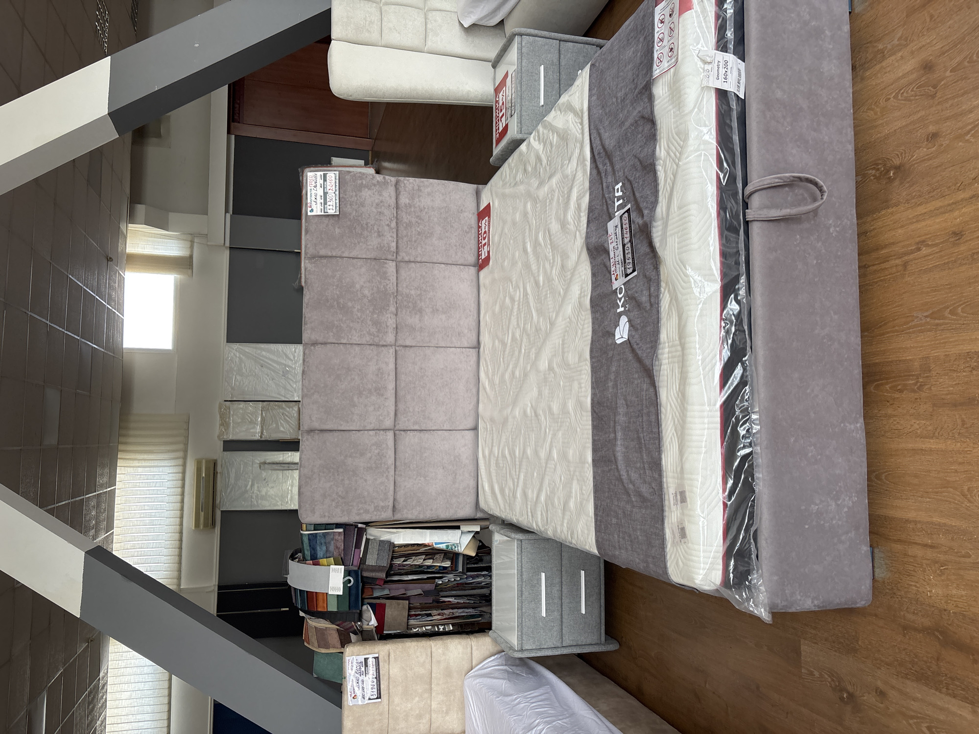 This is an indoor photograph of a furniture store, likely in Zaporizhzhia, Ukraine, showcasing mattresses and furniture. The scene is dominated by large, upholstered items, including several mattresses laid out horizontally and a modular sofa in a light grey color. One mattress is encased in clear plastic packaging, revealing a white quilted surface and a grey fabric band. Another mattress has a label with Cyrillic text and red markings indicating a discount.
In the background, there are stacks of what appear to be boxed furniture or mattress bases, some covered in plastic wrap. A collection of fabric swatches, organized in a binder, is visible on a lower shelf, suggesting a custom order or upholstery selection area. A narrow window with blinds lets in natural light, casting a soft glow on the scene. The floor is a warm wooden laminate.
Several price tags and labels are visible on the products. One tag on a mattress features Cyrillic text and the numbers "13300" and "30000". Another label on a mattress has "Geometry 160x200" printed on it. A small grey fabric box with silver accents is placed on top of another item. The overall impression is a well-organized retail space focused on bedroom furniture. There are no people visible in the image.