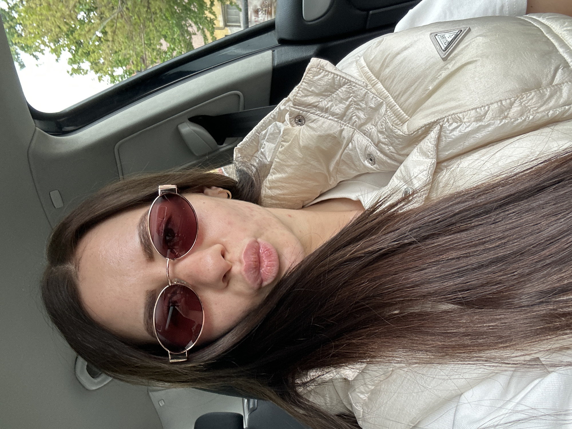 A young woman with long, brown hair and brown eyes is photographed indoors, seemingly in a car, with a view of green trees and a building visible through the vehicle's window. She is wearing sunglasses with a copper-colored frame and round, rose-tinted lenses, and a puffy, beige jacket with a triangular emblem on the left side. Her lips are pursed in a kissing gesture. The scene suggests a casual setting, possibly during transit. The exact location within Plovdiv, Bulgaria is indiscernible without further context.