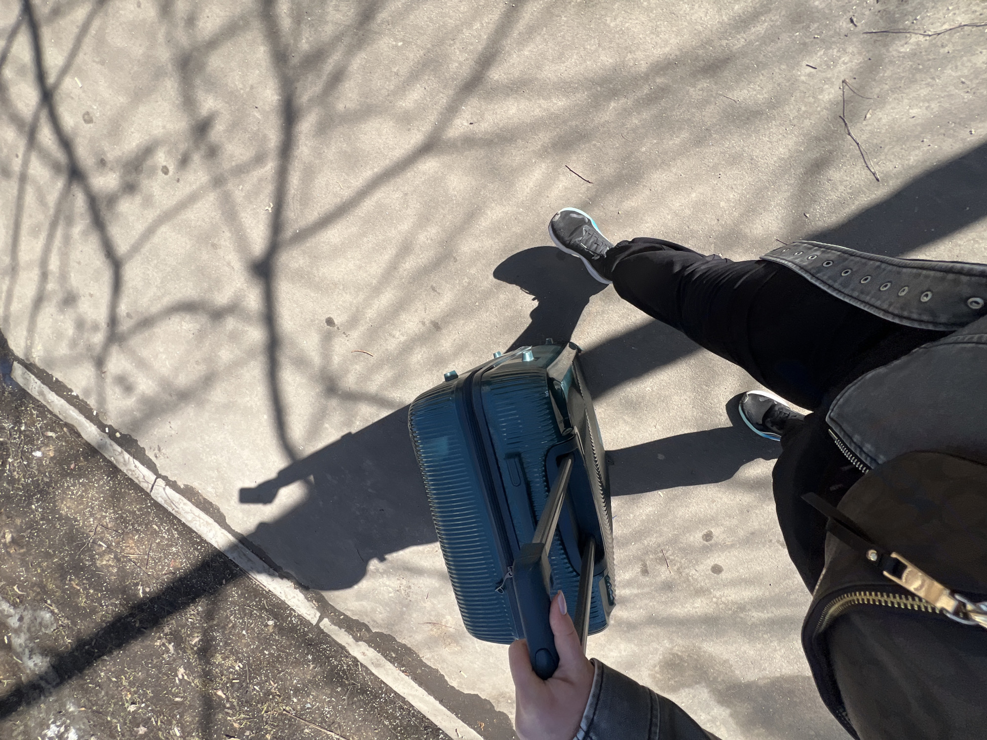 The image shows a first-person perspective of someone standing on a concrete surface, likely a sidewalk. The person is holding a teal-colored suitcase with a retractable handle. The person is wearing black pants and a black jacket with silver zippers and eyelets. Their feet are visible, wearing black and grey sneakers. The ground is partially covered in the shadows of trees. To the left of the person is a narrow strip of ground covered with what appears to be grass, dirt, and scattered debris. The location could be Moscow, Russia, as that information was requested in the prompt.