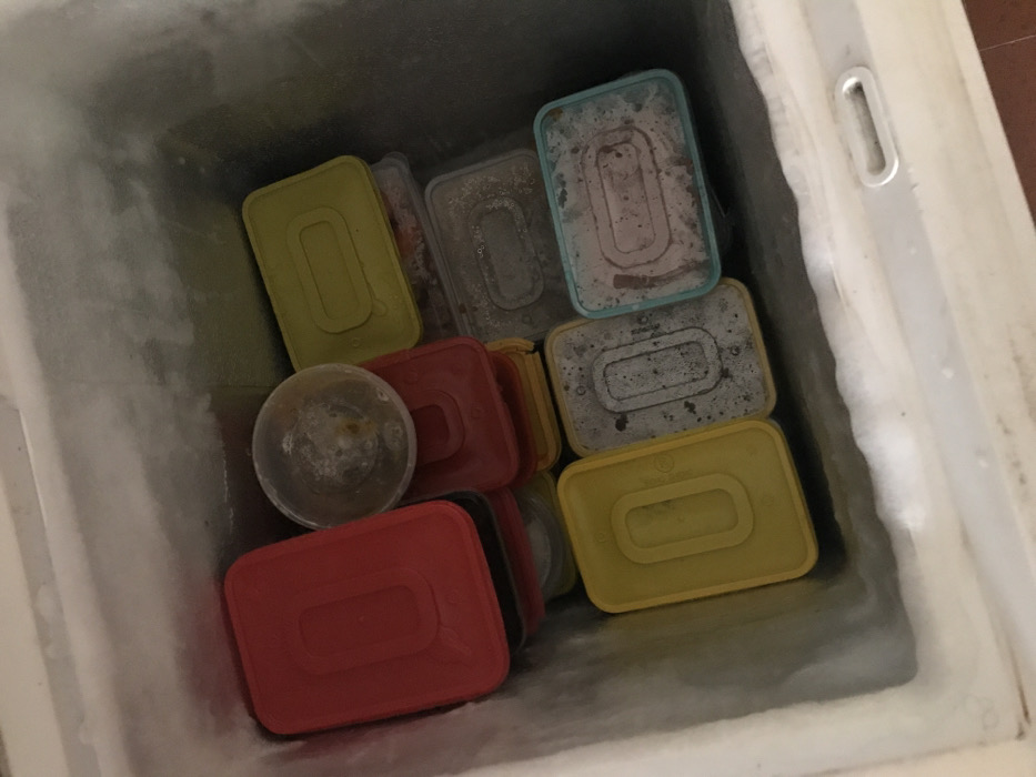 The image displays the interior of a chest freezer, viewed from directly above. The freezer's internal walls are heavily coated with a thick layer of white frost. The space is densely occupied by various plastic food storage containers, all appearing frozen.
Identified containers include:
- Multiple rectangular containers with opaque red lids.
- Multiple rectangular containers with opaque yellow lids.
- Several rectangular containers made of clear or translucent plastic, many showing significant frost accumulation on their surfaces. One of these translucent containers features a light blue rim.
- A single small, round, clear plastic container, also heavily frosted, positioned near the upper-center of the arrangement.
The contents of the containers are largely obscured by their lids or layers of frost, although some appear to contain dark or reddish food items. No individuals are present in the frame. The scene is located in Calabar, Nigeria.