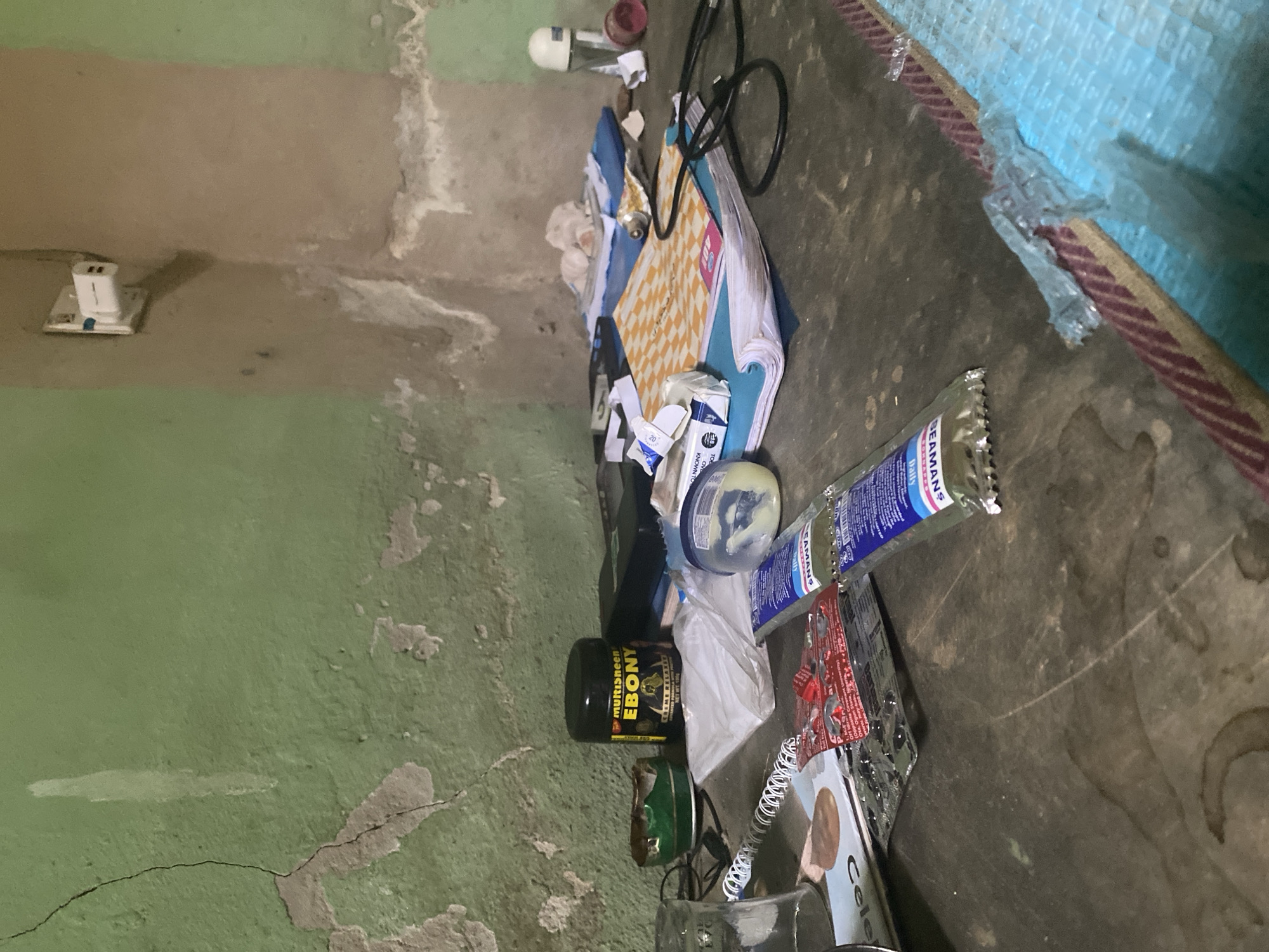 The image shows a cluttered corner of a room, with walls painted in green and brown tones with signs of wear and cracking. The floor is covered with a variety of discarded items including empty containers of hair products (e.g., "Ebony Creme Relaxer," "BEAMANS"), a notebook with a checkered pattern, electronic cables, packaging material, and a white wall plug. The scene is unkempt.