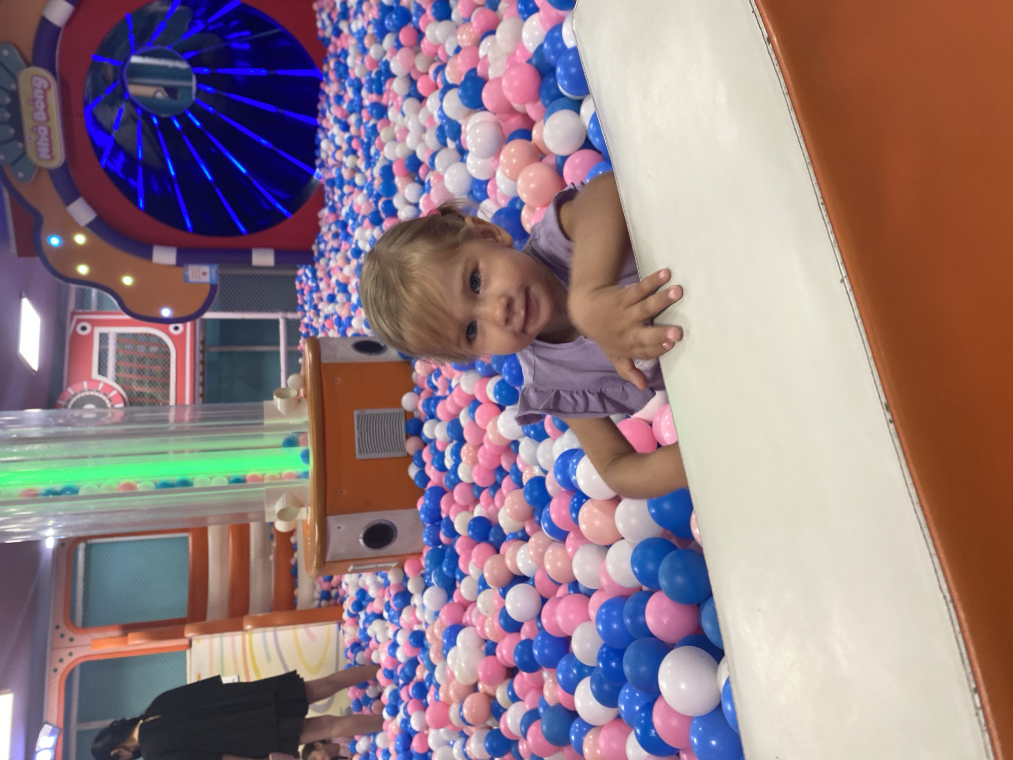 A young Caucasian girl with blonde hair and blue eyes is pictured in a ball pit, leaning on the edge of a white ramp. She is wearing a purple sleeveless top and has her left hand resting on the ramp. The ball pit is filled with blue, pink, and white plastic balls, creating a colorful environment. In the background, a large orange structure with a blue, circular, illuminated opening and a green transparent tube with balls are visible, suggesting an indoor playground. A woman in a black dress stands further in the background, on a set of orange stairs. The scene appears to be at a playground in the city of Phu Thuy, Vietnam.