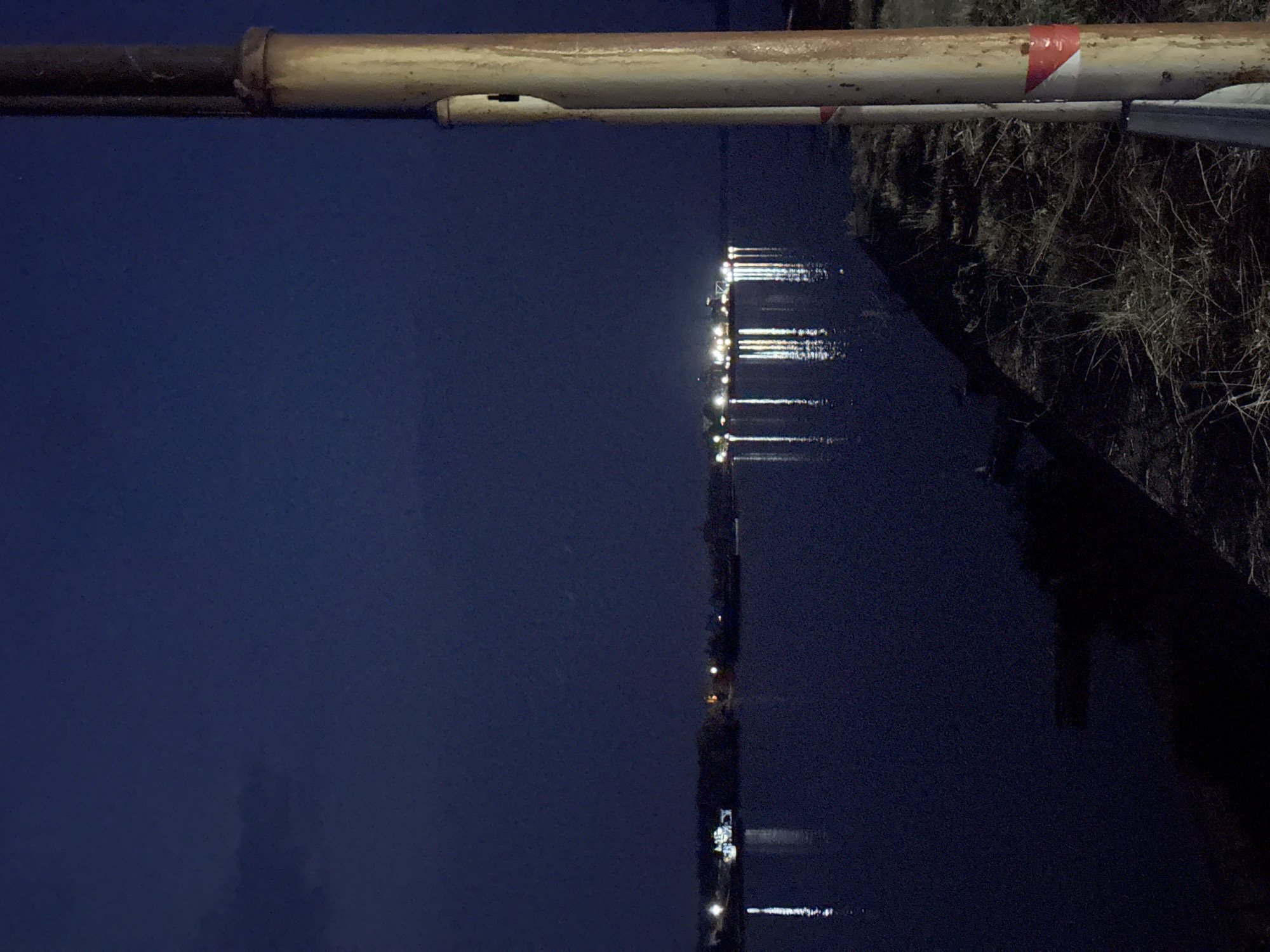 This is a nighttime photograph of a waterfront scene in the city of Tutrakan, Bulgaria. In the foreground, a rustic, weathered metal pole stands vertically on the right side of the image, showing signs of age and a white and red reflective band near its base. The pole is positioned on a grassy embankment that slopes down towards a dark body of water, likely the Danube River. The water is calm, with visible reflections of distant lights.
Across the water, a distant shoreline is illuminated by a cluster of lights, suggesting a settlement or industrial area. The lights create shimmering reflections on the water's surface, forming vertical streaks of light. The sky is a deep, dark blue, indicating the late hour, with a faint cloud formation visible on the left. The overall atmosphere is tranquil and nocturnal, highlighting the interplay of artificial lights and the natural darkness.