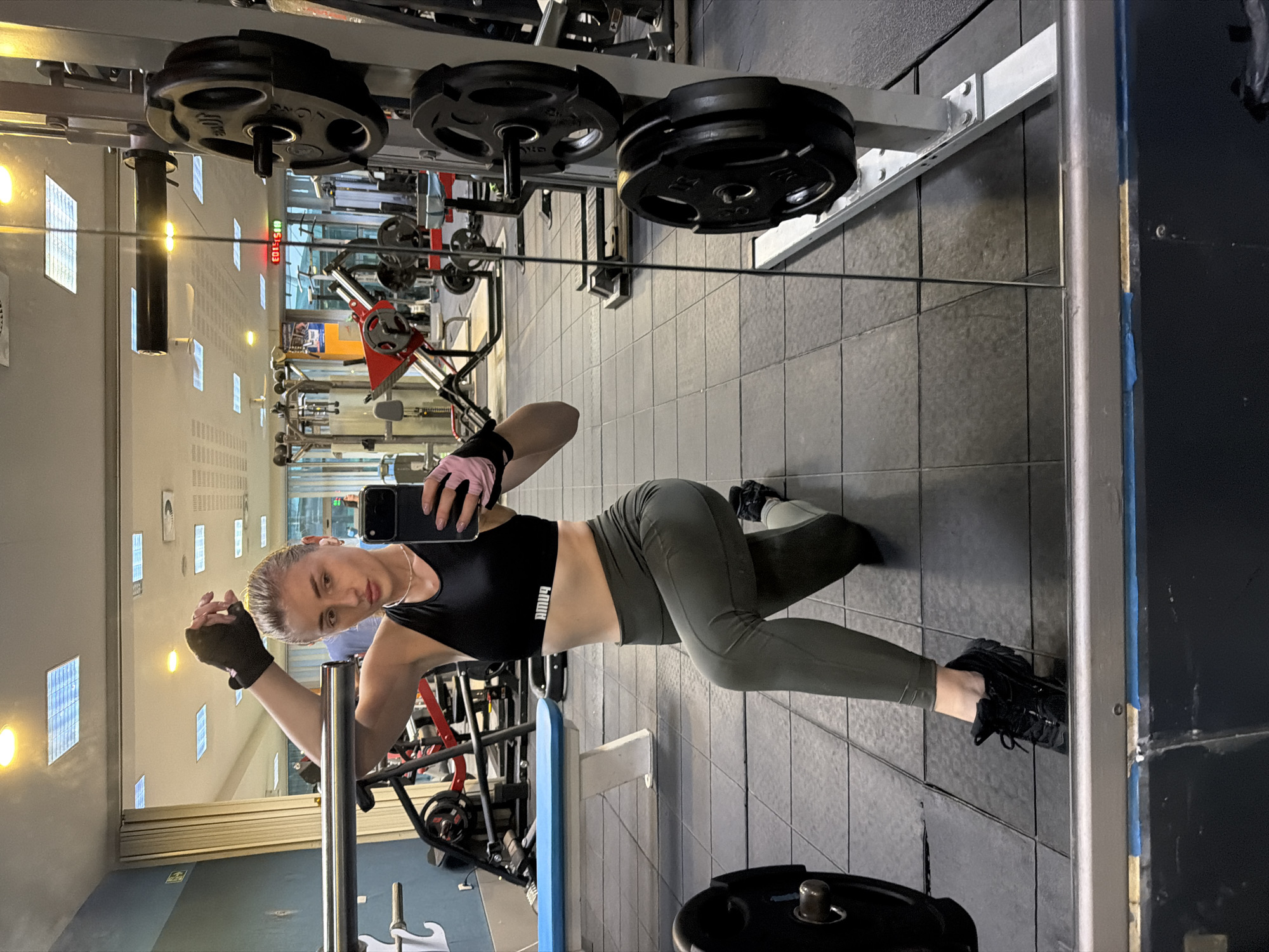 A woman is taking a mirror selfie in a gym. She is wearing a black sports bra and olive green leggings, black sneakers, and black and pink workout gloves. She is in a lunge position with her right knee on the floor. She is holding a black smartphone in her right hand, angled to capture her reflection. The gym is visible in the background, with weight racks, barbells, and other exercise equipment. The floor is tiled with black squares. The overall lighting suggests indoor artificial lighting.