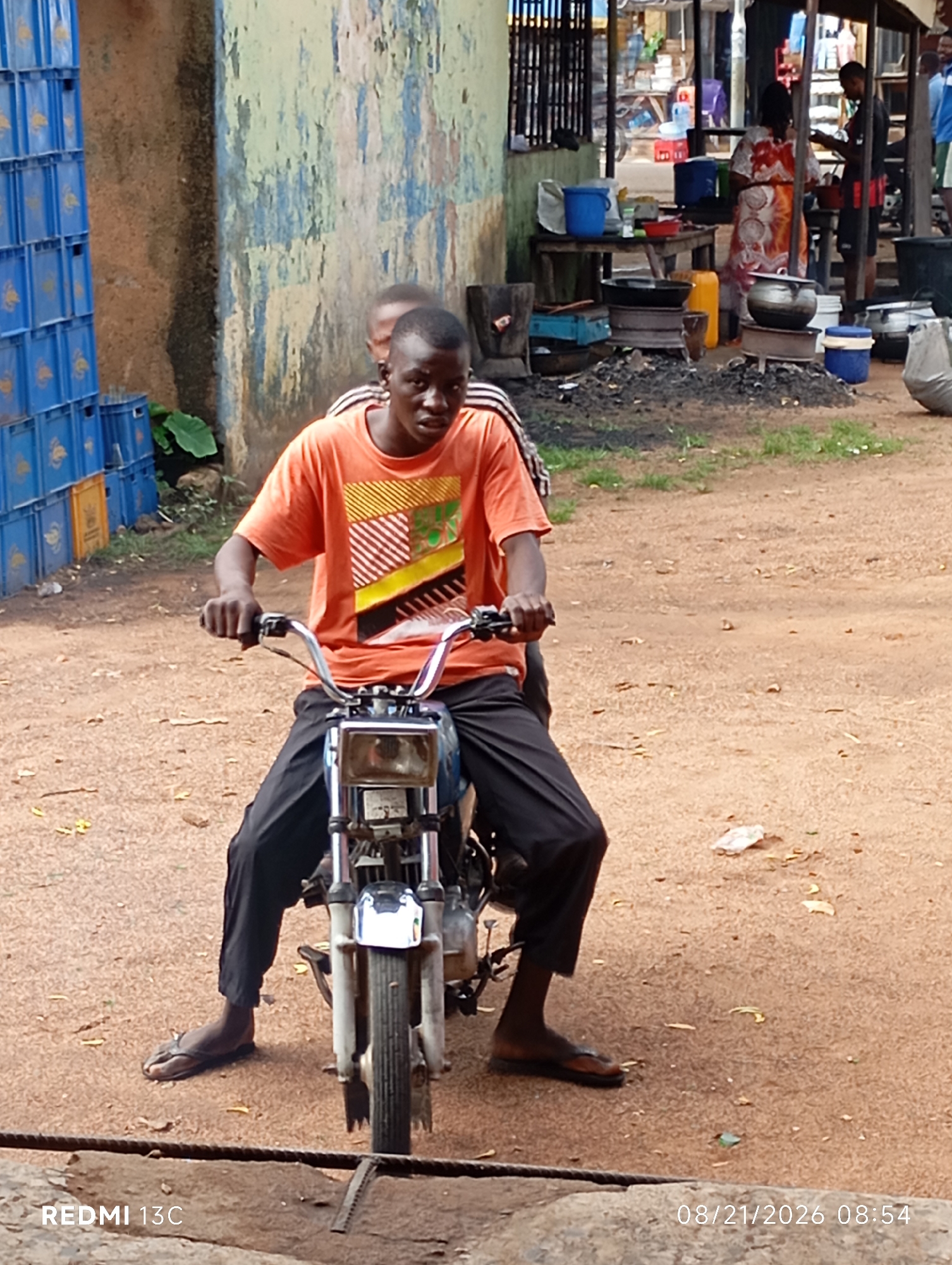 Two young Black males are on a motorcycle on a dirt road. The driver, wearing an orange t-shirt with a geometric design, is in the foreground. He is looking forward, holding the handlebars. A second young male, only his head and shoulders are visible, is sitting behind him. The motorcycle has a silver handlebar, a silver headlight, and is black with some silver accents. In the background, to the left, are stacked blue crates. To the right, there is a market stall with people and cooking equipment, including pots and a charcoal grill. The ground is a reddish-brown dirt with some scattered leaves and debris. A portion of a stone wall with a rope is in the lower foreground. A timestamp in the lower right corner reads "08/21/2026 08:54". The overall setting appears to be an outdoor market in a semi-urban or rural area.