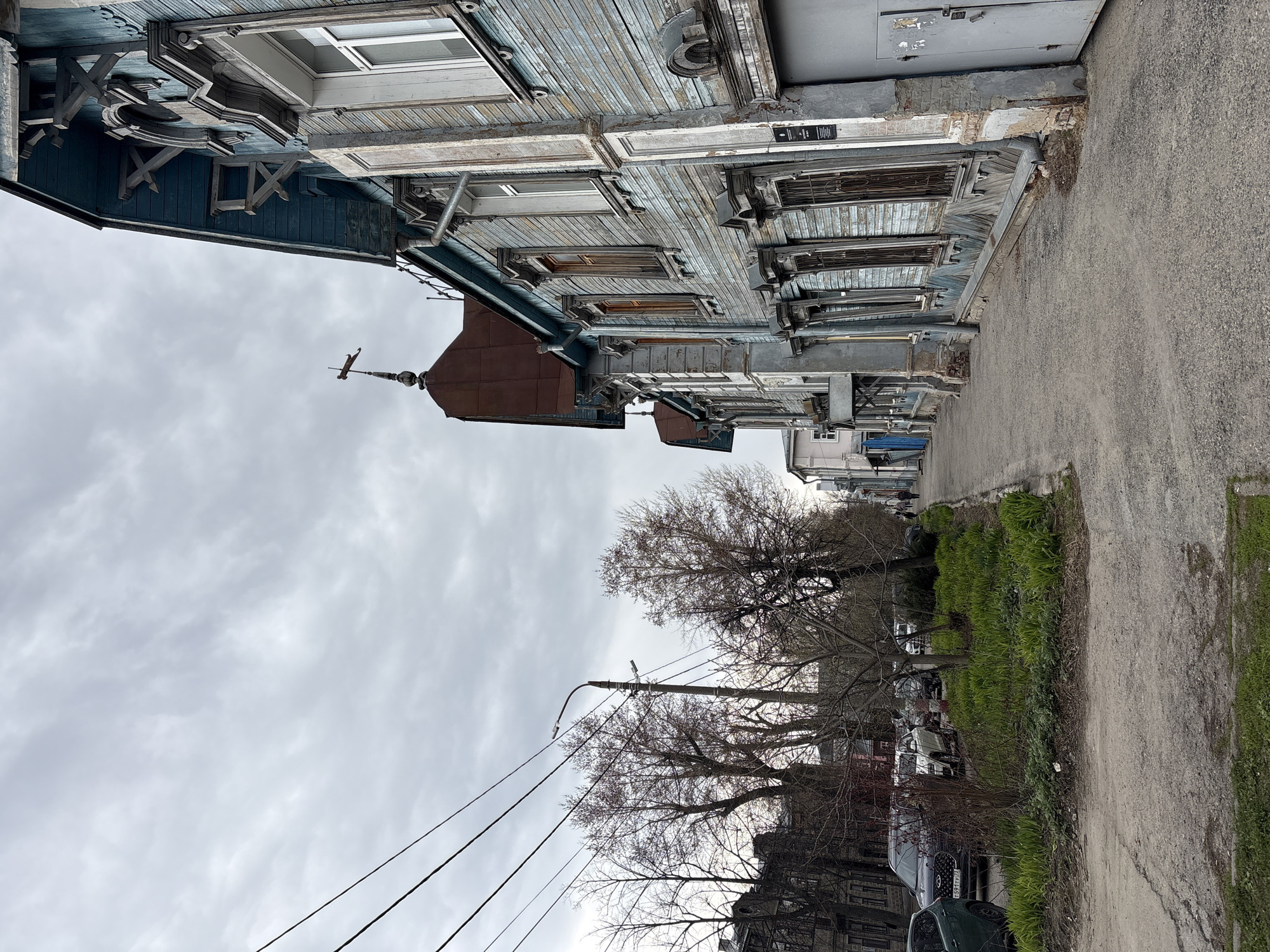 The image captures a street scene in what appears to be late winter or early spring, under an overcast sky. The primary subject is an old, two-story wooden building on the left, characterized by its faded blue/teal paint and extensive, intricate wooden fretwork and carvings, typical of traditional Russian architecture often found in cities like Ulyanovsk. The building shows significant signs of age and wear, with peeling paint and weathered wood on its facade and window frames. Many windows are visible, some with decorative surrounds, and others appear boarded or shuttered. A small, rust-colored dome with a weather vane sits atop a section of the roof.
Parallel to the wooden building runs a paved or asphalt road. Between the road and the buildings, there's a strip of ground covered with emerging green grass and bare deciduous trees, suggesting the onset of spring. Several cars are parked along the side of the road behind the trees and vegetation. A utility pole with power lines extends into the cloudy sky.
No specific activity is taking place; the scene is static, depicting a quiet street. The time of day is daytime, but the heavy cloud cover gives the light a soft, diffused quality.
Visible text includes a small, dark plaque on the facade of the blue wooden building, though its specific text is illegible. Another small sign, possibly for a utility, shows "ТЕЛЕФОН" (Telephone) and some numbers. A license plate on one of the parked cars clearly displays "797", which is a regional code used in Russia.