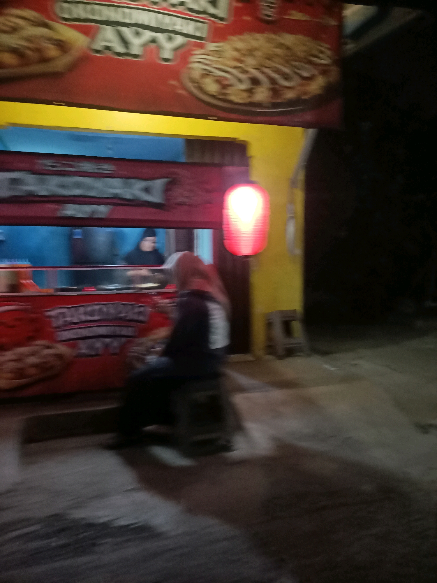 In Telagaasih, Indonesia, a food stall is open for business at night. The stall features a vibrant red awning displaying images of food, likely pizza. A red lantern hangs from the yellow wall of the structure, casting a warm glow. Inside the stall, an employee wearing a dark head covering can be seen preparing food. A customer, a woman in a hijab, sits on a stool in front of the stall, facing away from the camera. The ground in front of the stall is paved with concrete, and shadows are cast from the overhead lighting. The overall impression is of a lively, local eatery operating in the evening.