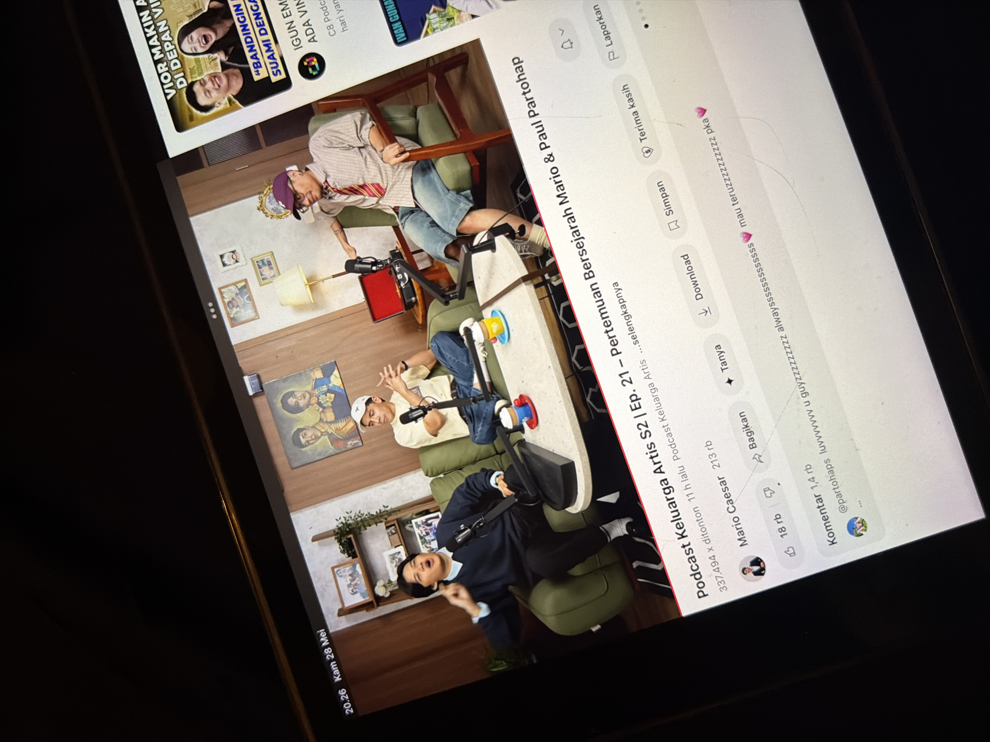 The image shows a screen displaying a podcast episode titled "Podcast Keluarga Artis S2 | Ep. 21 – Pertemuan Bersejarah Mario & Paul Partohap." The episode has been watched 337,494 times, with 11 hours since uploaded, and has 1.4 rb of comments. The episode is from Mario Caesar and had 213rb likes and 18rb shares. The screen also shows options like Tanya, Bagikan, Download, Simpan, and Terima kasih, and Laporkan. The broadcast is located in East Jakarta, Indonesia. The people on the screen are seated on sofas in a studio setting, with a coffee table in front of them. The backdrop consists of wood paneling, framed pictures, and plants. The environment is dimly lit.