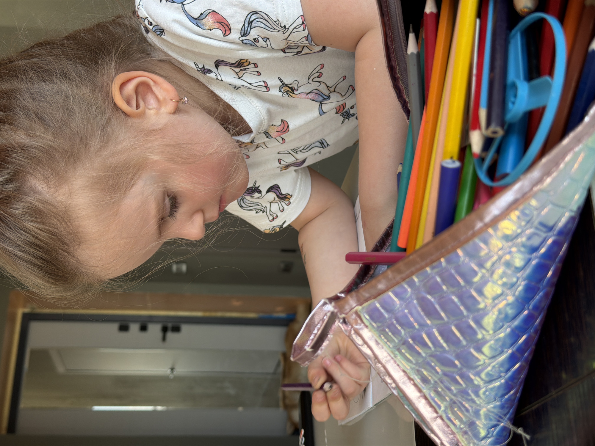 A young girl with blonde hair is shown in profile, looking down and drawing. She is wearing a white t-shirt with a unicorn pattern. Next to her is a pencil case filled with colored pencils and a blue pencil sharpener. The background includes a light-colored door. It is not possible to determine the location, Chaikovsky, Russia, based on the visual information in the image.