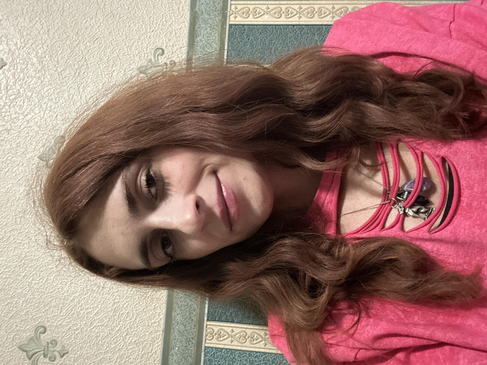 The media file is a portrait of a young woman with long, wavy, auburn hair. She is wearing a bright pink shirt with cutouts across the chest, revealing a necklace with a silver pendant. Her face is turned towards the camera, and she has dark eyes, eyebrows, and mascara. Her lips are slightly parted, and she appears to be smiling faintly. The background consists of textured wallpaper in muted tones and a decorative border with a repeating pattern. The lighting suggests an indoor setting, possibly during the daytime. The image has a vertical orientation. No visible text is present.