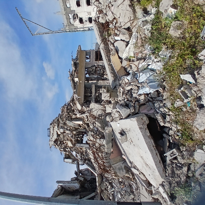 This image presents a scene of extensive structural destruction in Jabalia, Palestinian Territory. The foreground and midground are dominated by a large mound of rubble, consisting primarily of fractured concrete slabs, broken blocks, and twisted steel rebar. This debris appears to be the remnants of collapsed multi-story buildings. In the center-midground, a partial structure with exposed internal elements and empty window or door frames remains somewhat intact amidst the surrounding wreckage. To the far left, a grey utility pole stands vertically. To the right, a partially damaged, multi-story concrete building with numerous dark, empty window openings is visible in the background, alongside a tall, skeletal metal framework that resembles a structural support or pole. The ground in the immediate foreground shows sparse patches of green vegetation growing through the rubble. The sky above is clear blue with minimal scattered white clouds. No individuals are present in the frame.