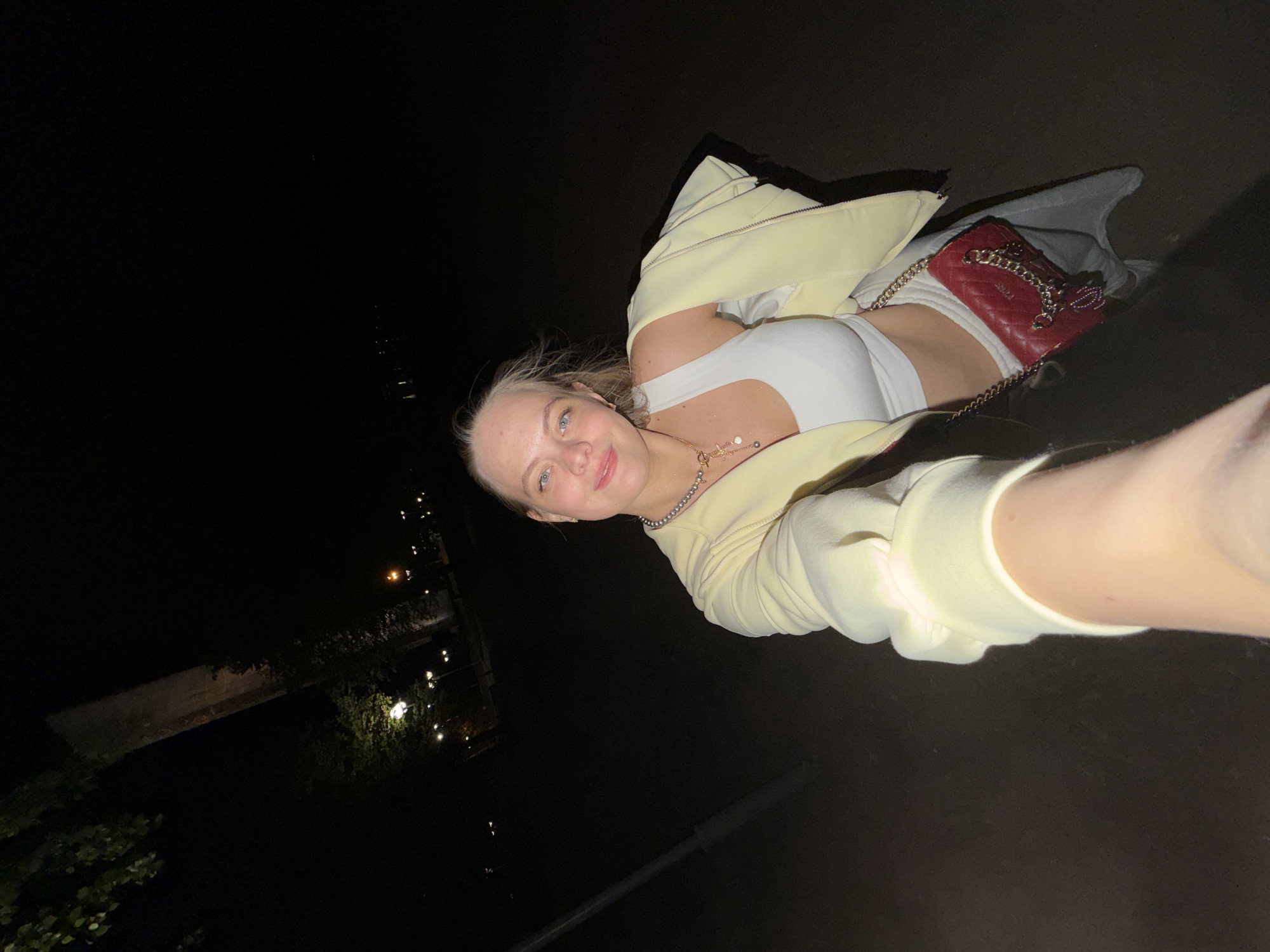 At night, a young Caucasian woman poses for a selfie in Izhevsk, Russia. She wears a white crop top, pale yellow jacket, and necklace. A red purse rests near her midsection. Streetlights illuminate a dark urban landscape.