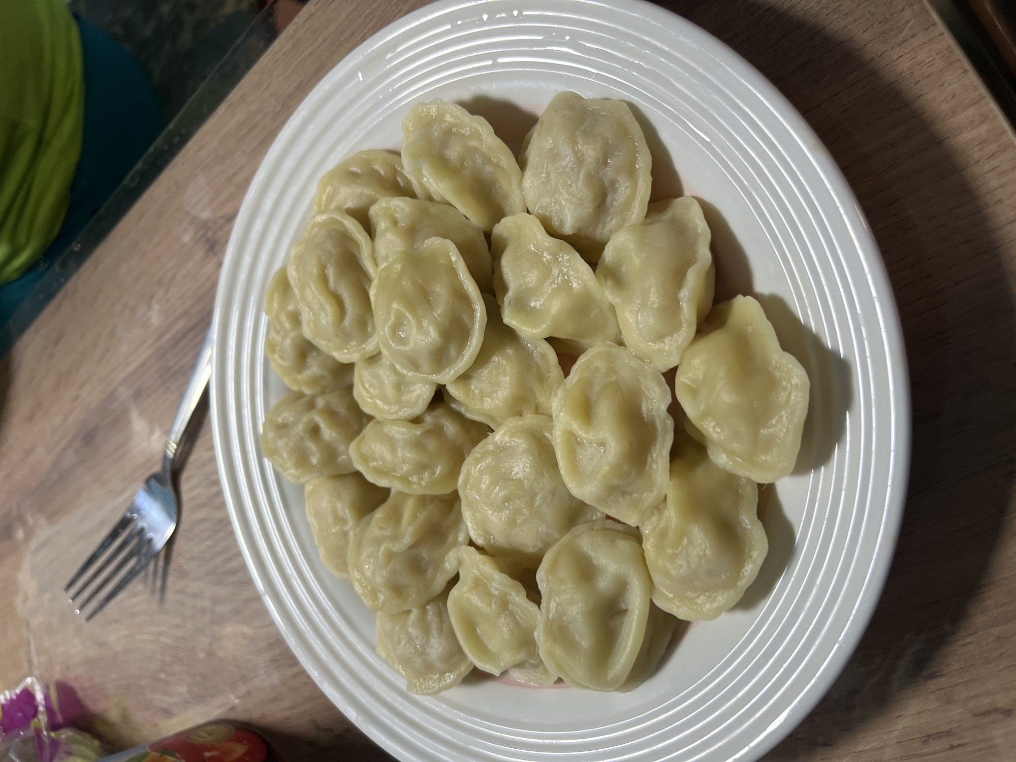 A plate of boiled dumplings is presented on a wooden surface. The dumplings are arranged in multiple layers, filling most of the white plate. A fork rests on the table to the left of the plate. In the background, a glimpse of a person wearing a green shirt and a blurry container can be seen. The scene appears to be indoors, possibly a kitchen or dining area. The lighting suggests it is daytime. This image does not provide any information related to the city of Nizhny Tagil, Russia.