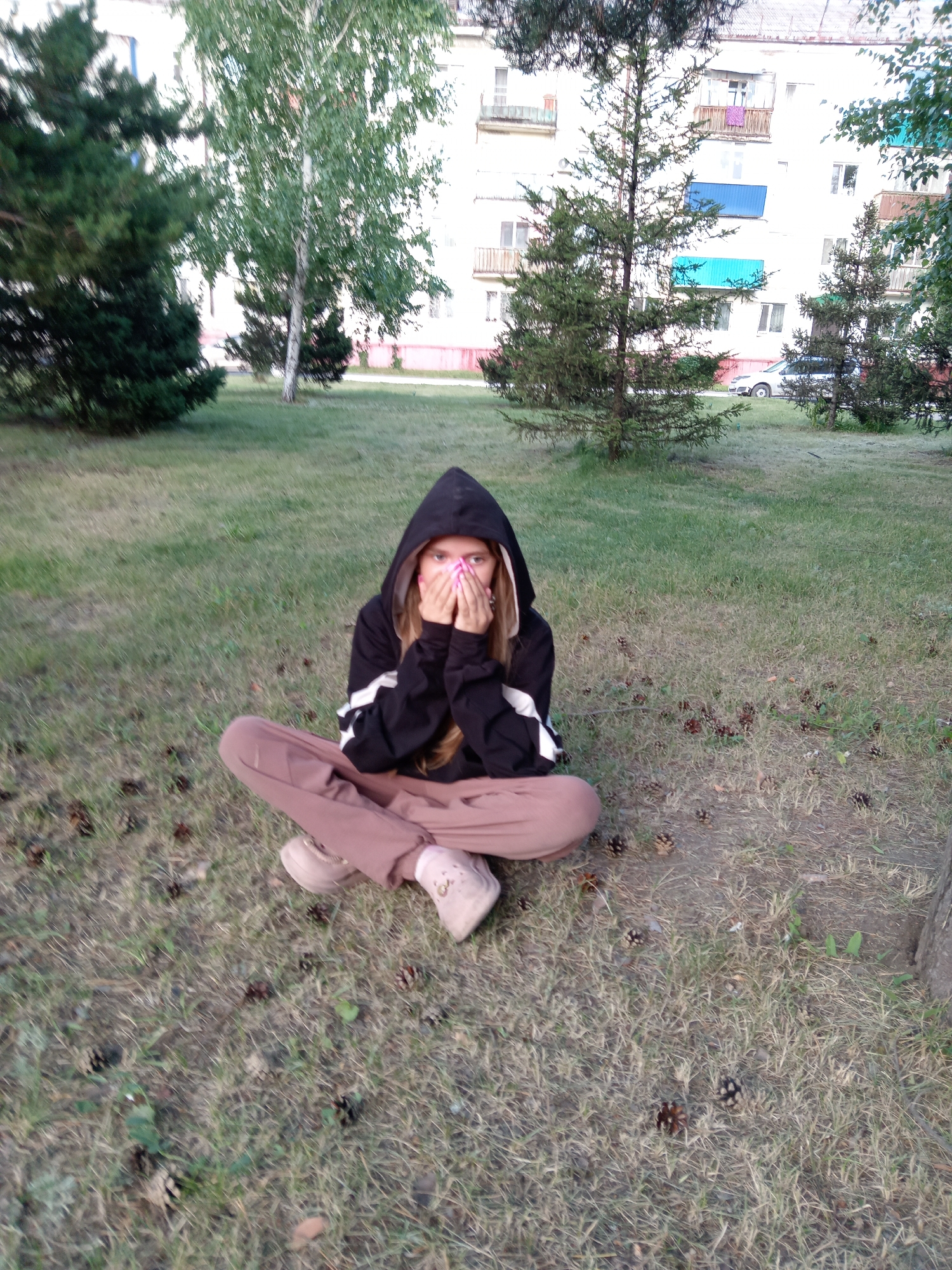 A young female is sitting cross-legged on a grassy lawn, facing the camera. She is wearing a black hooded sweatshirt with white stripes on the sleeves and light brown pants. Her hands are covering her mouth, and she appears to be hiding her face. The background shows a multi-story apartment building, some trees, and a car parked in front of the building. The scene is outdoors, likely in a residential area of Rudny, Kazakhstan. The ground is covered with dry grass and scattered pinecones.