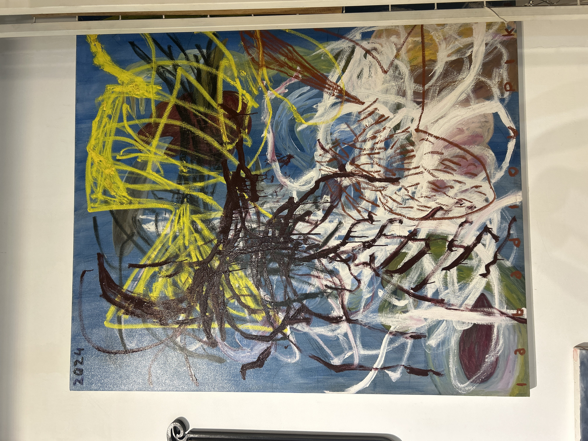 An abstract painting, rendered with expressive and dynamic brushstrokes, is presented against a blue background. The artwork features a complex interplay of lines and forms in multiple colors, including bright yellow, dark brown/maroon, black, white, and subtle hints of pale pink and green.
On the left side of the composition, vibrant yellow lines create a dense, tangled network, overlapping and extending towards the center. Over and beneath these, dark brown, maroon, and black strokes form more pronounced, almost organic, branch-like or root-like patterns that crisscross the canvas, particularly in the central and lower regions. Towards the right, broad, sweeping white strokes swirl and overlap, suggesting movement and depth, contrasting with the darker and more linear elements. The layers of paint create a textured appearance, indicating a gestural style.
The painting is mounted on a plain white wall, likely within a gallery or exhibition space, as suggested by the white track or railing visible along the top edge of the frame. A portion of another artwork or object is faintly visible on the lower right side of the overall scene.
Visible text on the painting includes the year "2024" written vertically in black in the bottom left corner. Along the right edge of the canvas, faint, vertically aligned characters or symbols can be discerned, possibly including "k", "D", and "o" or "p".