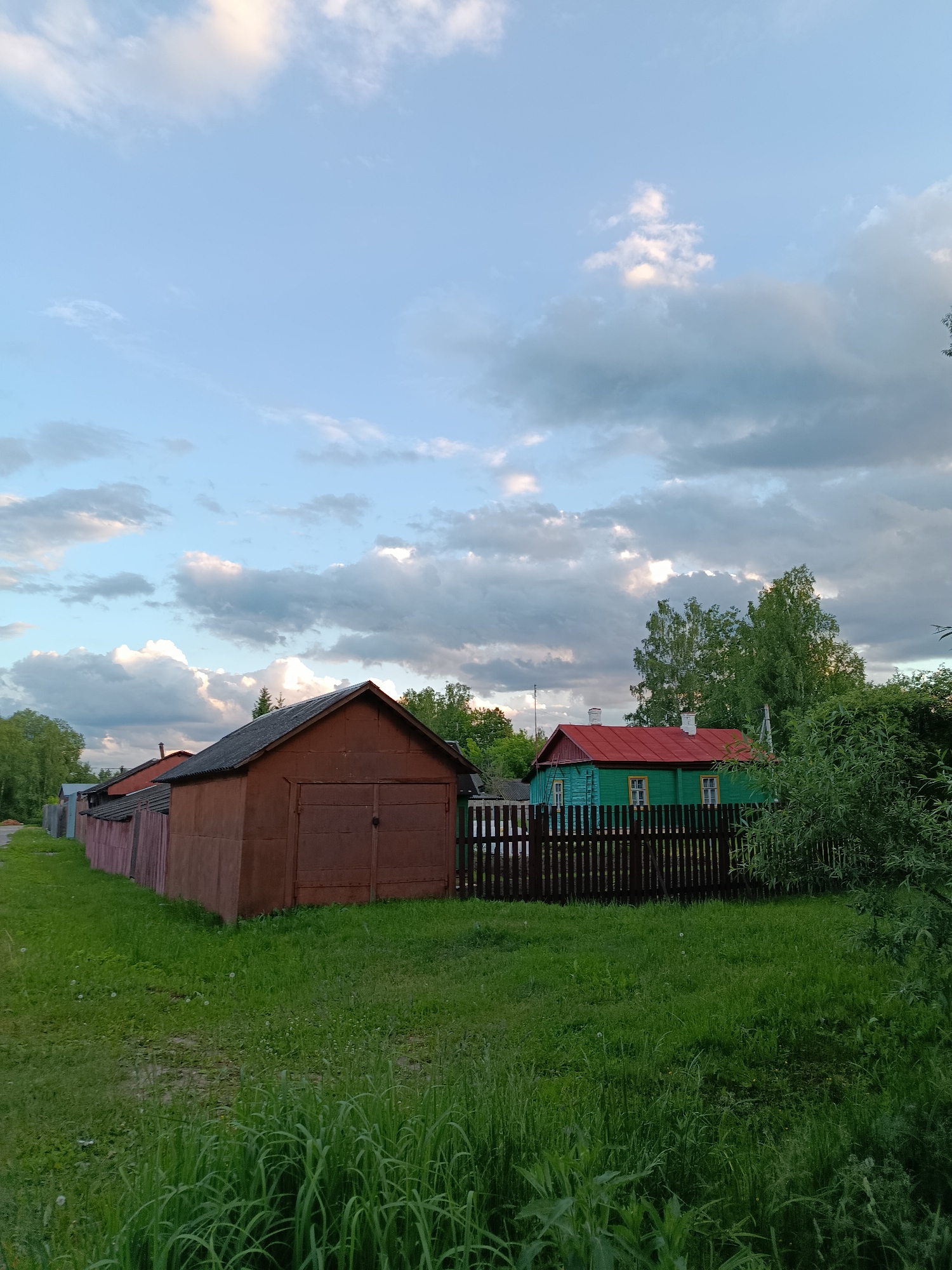 The image displays a tranquil village scene, likely set in Verea, Russia, under a partially cloudy sky. The time of day appears to be late afternoon or early evening, indicated by the soft, directional light illuminating patches of the clouds. The sky, a clear blue in parts, is adorned with numerous white and grey cumulus clouds.
In the midground on the left, a sturdy, dark reddish-brown wooden garage or shed with double doors is prominent. To its left, a long, lower wooden structure or fence, painted a faded reddish-pink, stretches along a dirt path or road. The immediate foreground is covered in lush, overgrown green grass and foliage.
On the right side of the image, behind a dark wooden picket fence, a vibrant green wooden house with a bright red roof and two white chimneys is partially visible. This house is surrounded by dense green trees and bushes, which also frame the right side of the photo.
The scene is devoid of people or discernible activities, conveying a peaceful and quiet atmosphere typical of a rural setting. There is no visible text in the image.