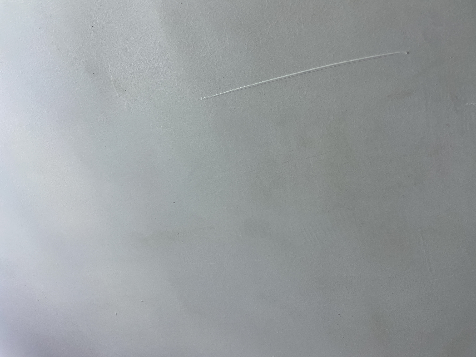 This is a close-up, top-down photograph of a textured, pale gray surface, possibly a wall or ceiling. A single, thin, diagonal scratch is visible, running from the upper left to the lower right of the frame. The scratch appears to be a superficial mark on the surface, with no indication of damage to a substrate. The lighting suggests an indoor setting, with the light source likely originating from above and to the left, casting subtle shadows that highlight the texture of the surface. There are no people, objects, or discernible location cues within the frame. The overall impression is one of a neutral, unremarkable surface. No text is visible in the image.