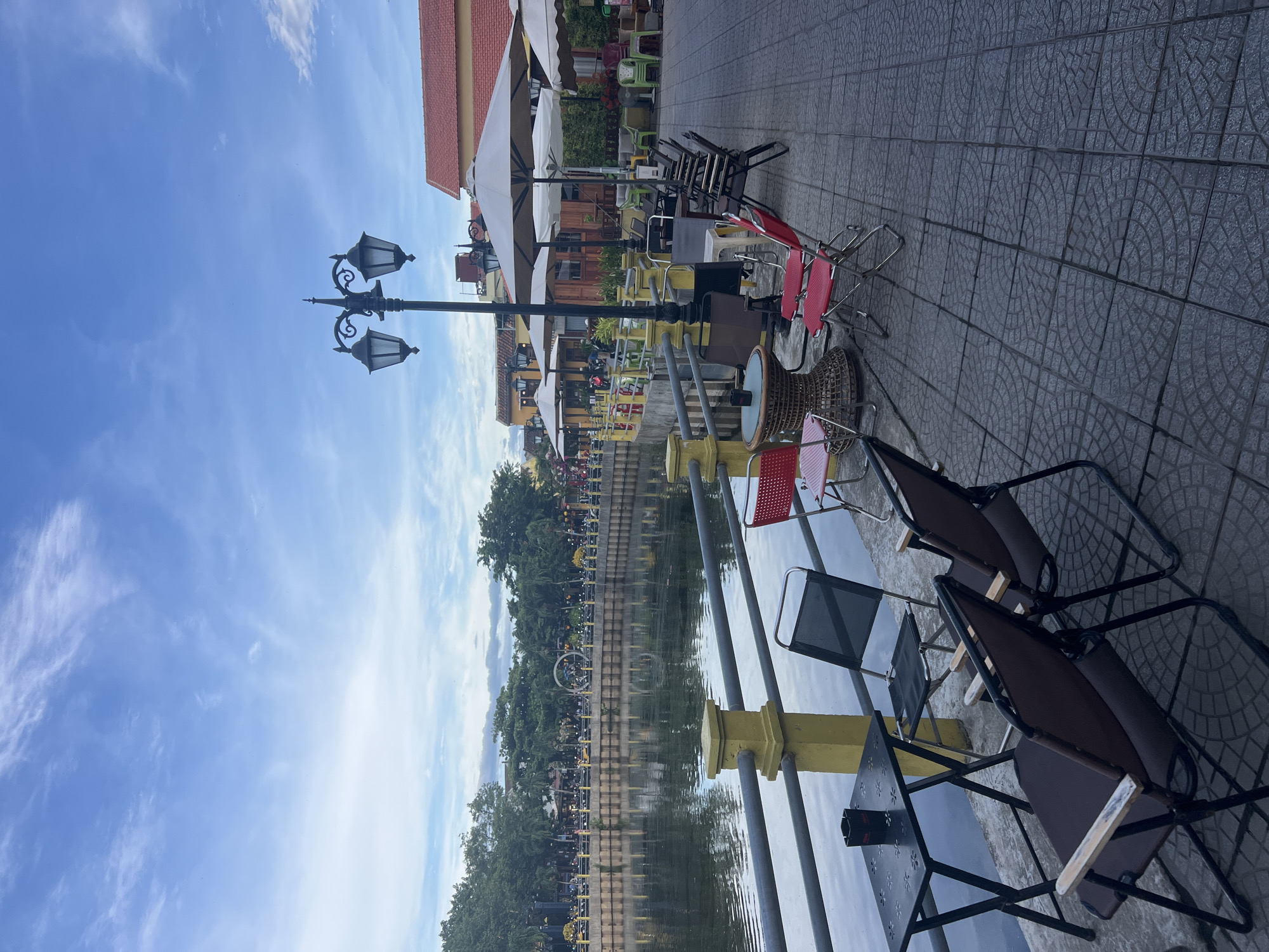 An outdoor cafe seating area is set up along the edge of a calm body of water, likely a canal or river, in Hoi An, Vietnam. The scene is captured from a slightly elevated perspective, looking towards the water and across to a tree-lined promenade. The sky above is a soft blue with wispy white clouds, suggesting it is late afternoon or early morning.
In the foreground, a black folding table with a textured surface is set up with an ashtray. Two brown folding chairs with wooden accents are positioned in front of the table, angled slightly towards the water. To the left of these chairs, a dark grey folding chair with a black fabric seat is placed next to the table.
Beyond the foreground seating, the tiled pavement continues, leading to more tables and chairs. Several red folding chairs are visible, along with a woven round table. Further back, white umbrellas are deployed over tables, indicating a covered seating area. A decorative lamppost with two ornate lanterns stands prominently near the cafe seating.
The body of water reflects the sky and the buildings on the opposite bank. Along the water's edge on the far side, a yellow railing runs the length of the promenade, separating it from the water. Behind the railing, a row of trees with dense foliage creates a lush green barrier. In the distance, buildings with red tiled roofs and yellow facades are visible, characteristic of Hoi An's architecture. People are present on the promenade across the water, with some activity suggested by the lights and shapes of vehicles and pedestrians. The overall impression is a tranquil, yet lively, waterside cafe environment.