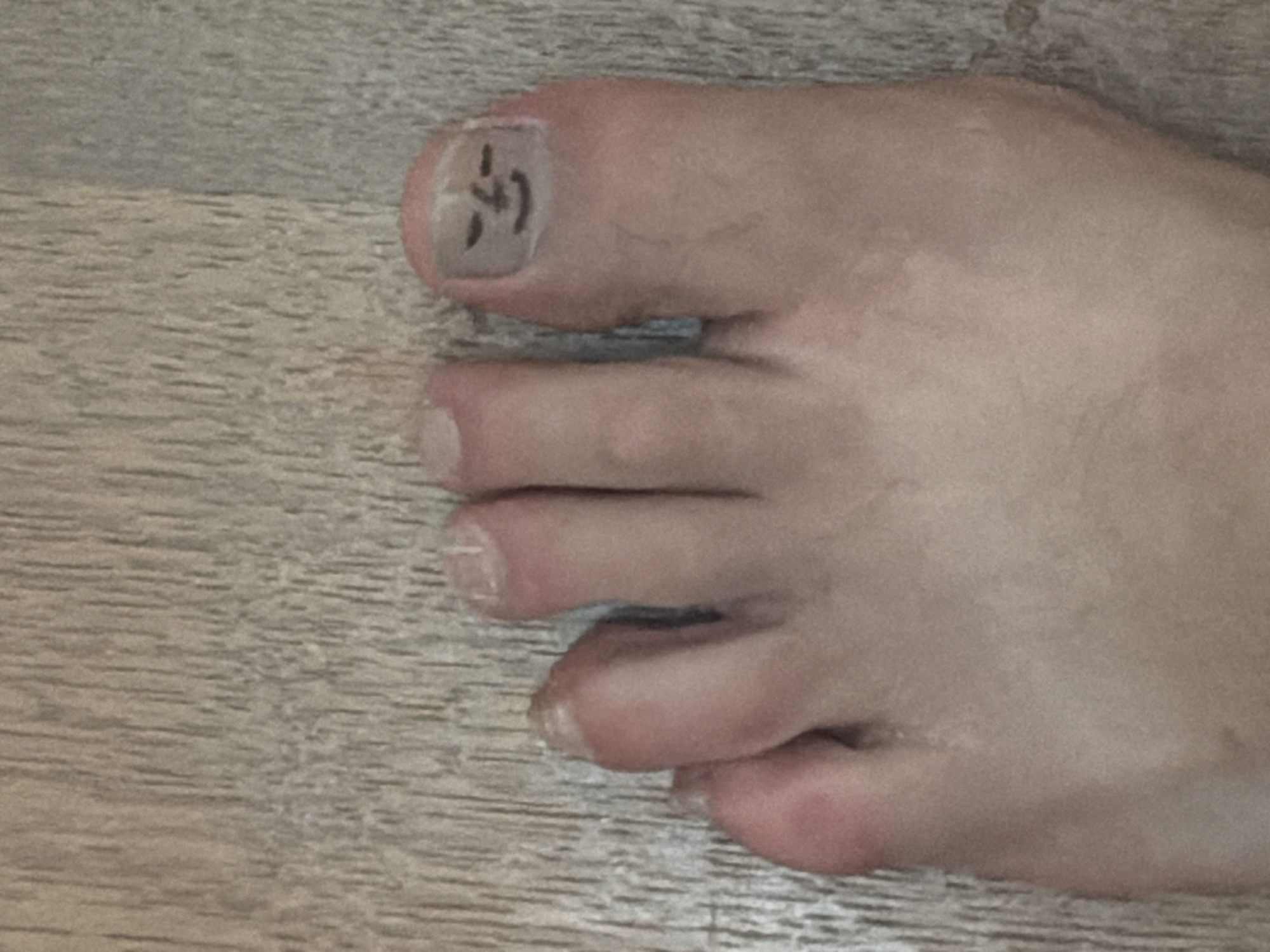 The image shows a close-up of a human foot with a drawing of a face on the toenail of the big toe. The foot is positioned on a wooden floor. The face on the toenail appears to be a simple drawing with eyes and a smiling mouth. The toes are slightly splayed. The wooden floor has a visible grain pattern and is light brown in color. There are no people or other significant objects in the scene. There is no information in the image to suggest its location in Esch-sur-Alzette, Luxembourg.