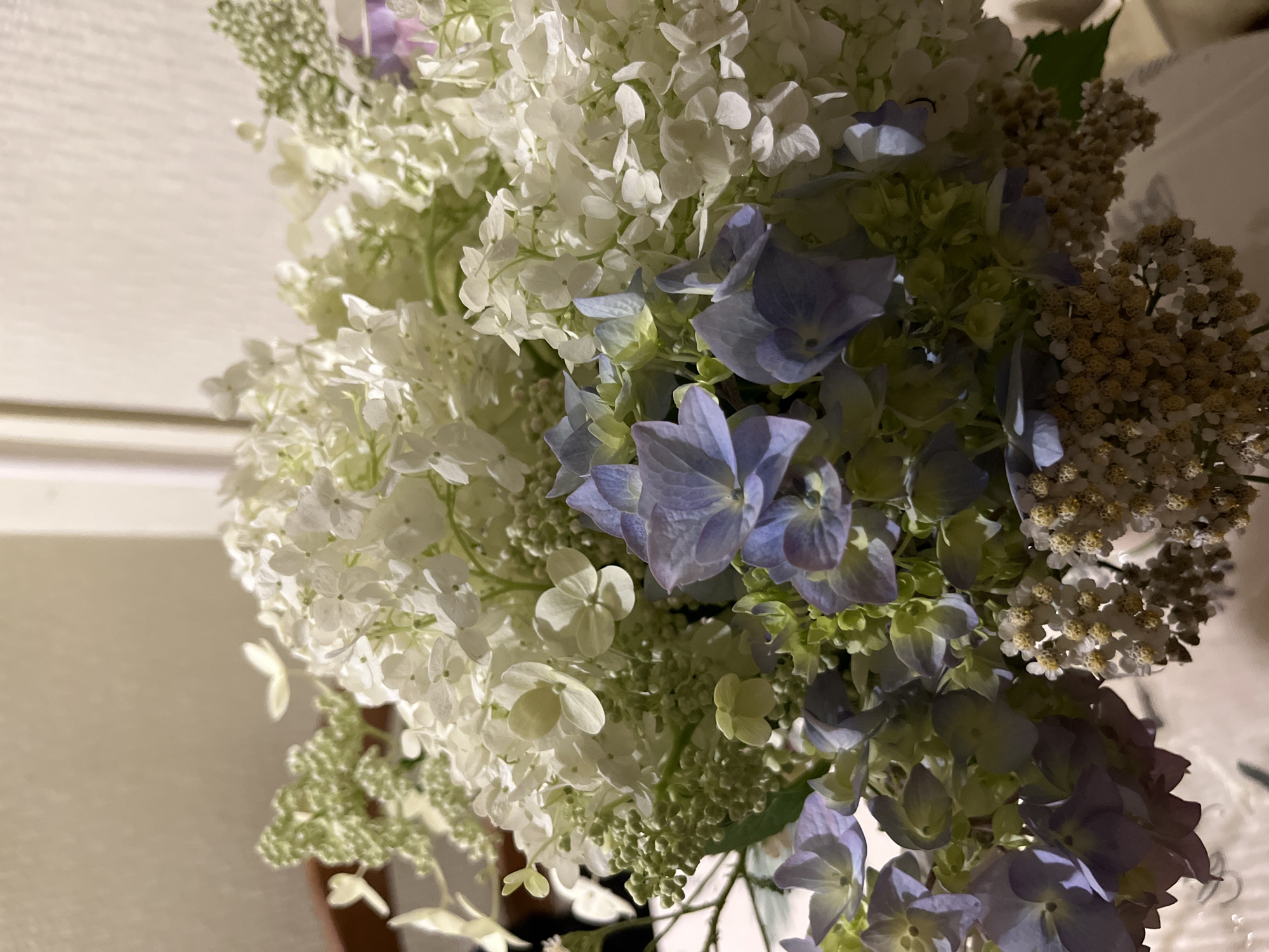 A bouquet of hydrangeas and other small white flowers is displayed in a vase. The hydrangeas are a mix of white and pale blue. The background is a neutral wall.