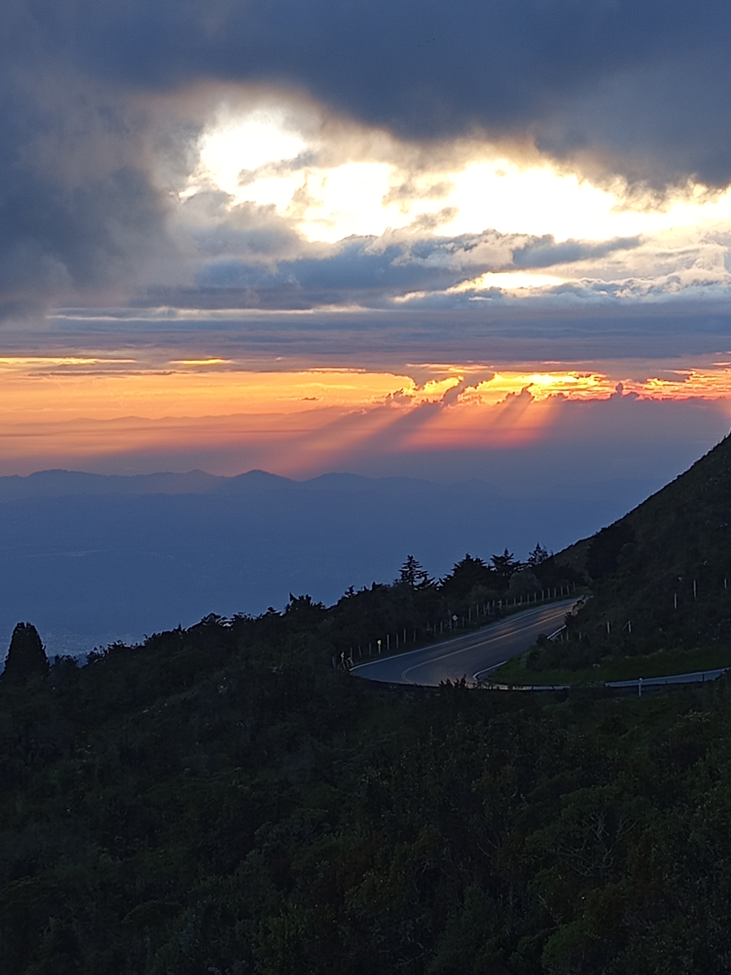 The image captures a dramatic sunset over a mountainous landscape. In the foreground, a dark, lush hillside with dense vegetation leads to a winding paved road that curves sharply. The road is illuminated by the fading light, with some streetlights visible along its edge. Beyond the immediate hillside, layers of hazy blue mountains recede into the distance. The sky is a spectacle of color, with deep grays and blues at the top transitioning to vibrant oranges and yellows where the sun is setting. Sunbeams radiate through gaps in the clouds, creating a striking visual effect. The overall impression is one of natural beauty and a serene, almost ethereal atmosphere. The location is identified as the mountainous area near Piedecuesta, Colombia, likely a viewpoint overlooking the city and its surrounding valleys. No people or specific actions are depicted in the image.