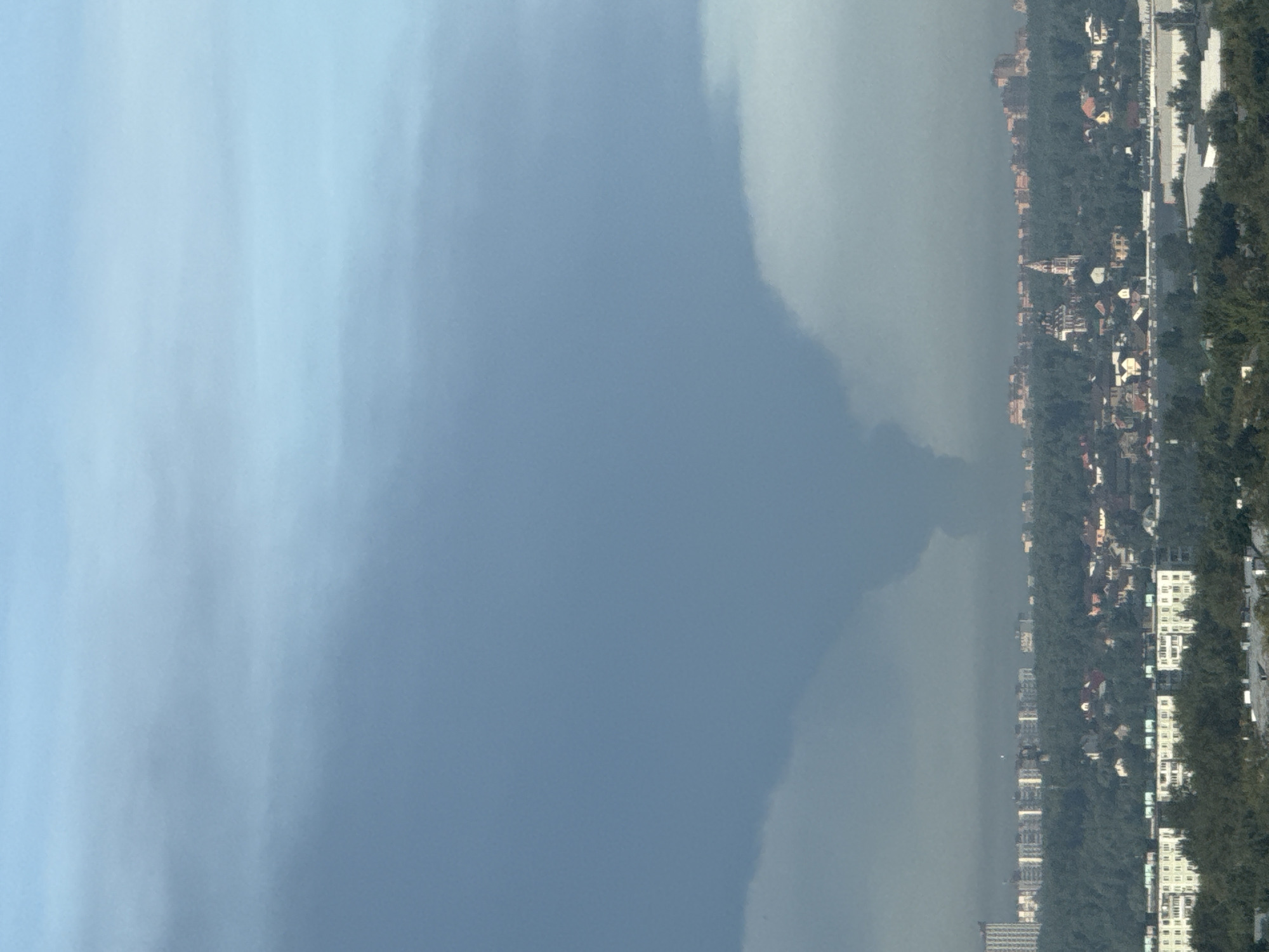 This image shows a wide shot of the city of Lyubertsy, Russia, under a dramatic sky. In the foreground, dense green trees partially obscure a collection of residential buildings, primarily apartment blocks. Behind these, a swath of mixed-use development is visible, including more apartment buildings and smaller houses nestled amongst trees. Further in the distance, a cluster of multi-story buildings can be seen, including a tall, modern high-rise on the left. The most striking feature of the image is the sky, which is dominated by a dark, funnel-shaped cloud formation, appearing to descend from the upper atmosphere towards the city. The lower part of this formation is darker and more defined, suggesting a turbulent weather event. The rest of the sky is a hazy blue, with lighter clouds visible. The overall impression is one of a looming weather phenomenon over an urban landscape.