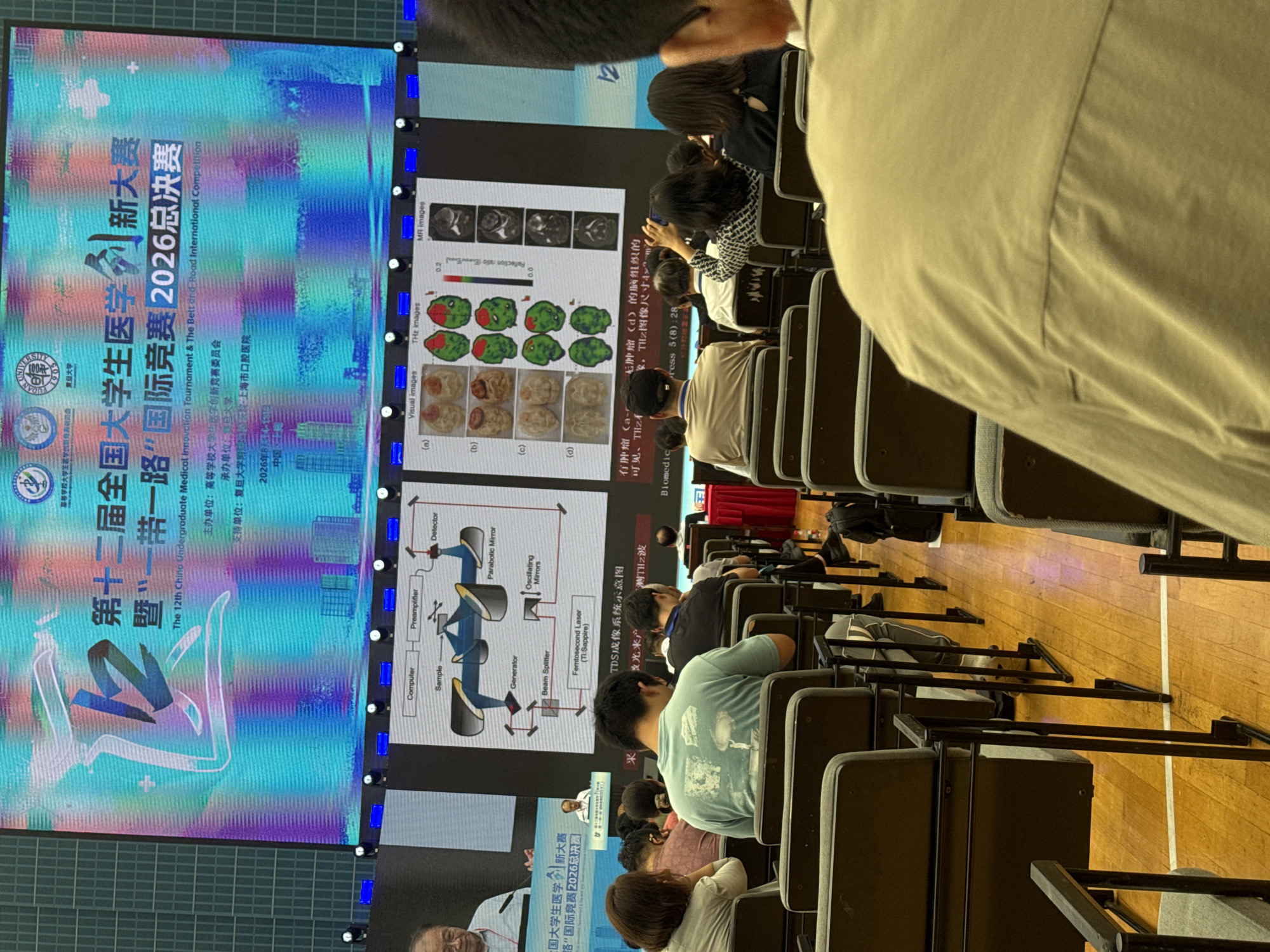 The image depicts a large screen displaying information related to an academic competition. The screen shows Chinese text and graphics, including a schematic diagram of a scientific instrument. Below the screen, an audience is seated in an auditorium, facing the screen. The seats are arranged in rows, and some audience members are visible from behind. The environment appears to be a conference hall or lecture theater. The presence of the large screen and audience suggests an event such as a presentation, competition, or lecture. The text on the screen indicates it is the "12th China Undergraduate Medical Innovation Tournament & The Belt and Road International Competition 2026 Finals." The location is identified as Yangpu District, China, based on the provided context.