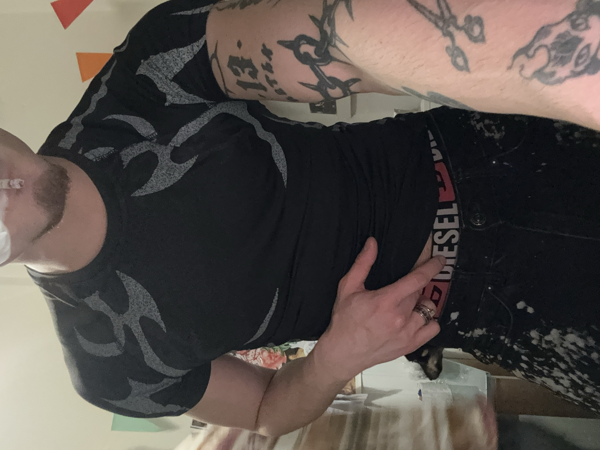 A man with tattoos on his arm and a goatee is wearing a black t-shirt with gray designs and black pants with white splatters. He is pointing at the word "Diesel" on his waistband. The background appears to be a room with a wall partially visible, featuring geometric shapes. The location is unknown and cannot be determined from the image, and no information suggests Sofia, Bulgaria.