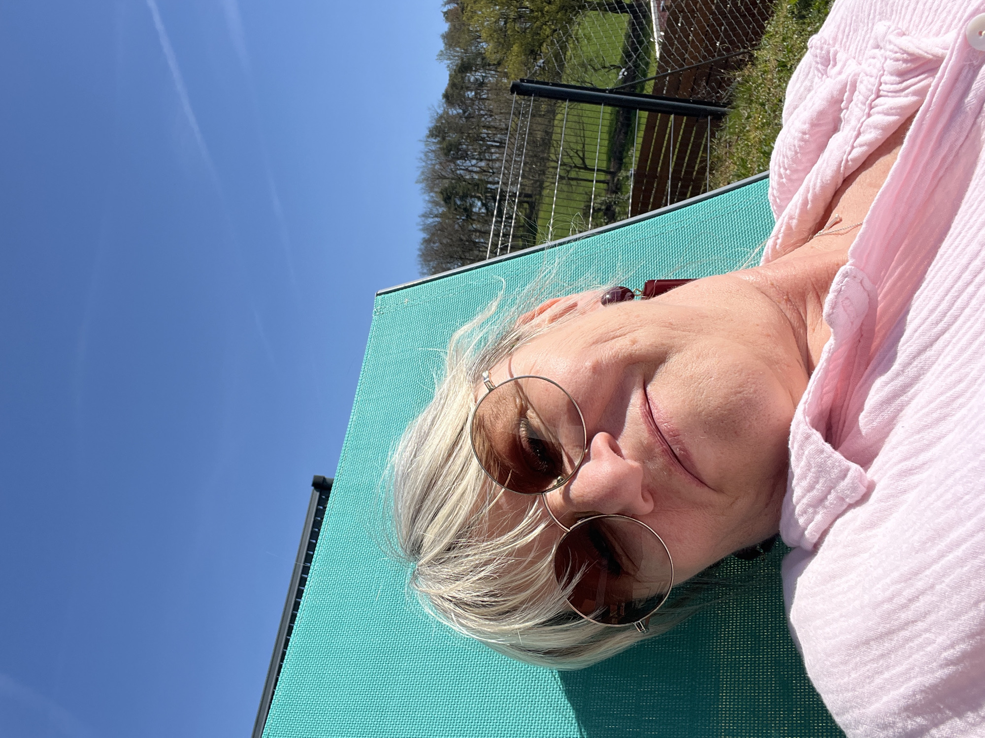 The image is taken outdoors, likely in a backyard in Chambœuf, France, as indicated by the location. A woman is lying on a turquoise lounger. She has short, blonde hair and is wearing round, brown sunglasses. She is dressed in a light pink, textured shirt. Behind her is a metal fence, a grassy area, and a treeline. The sky is clear and blue with contrails.