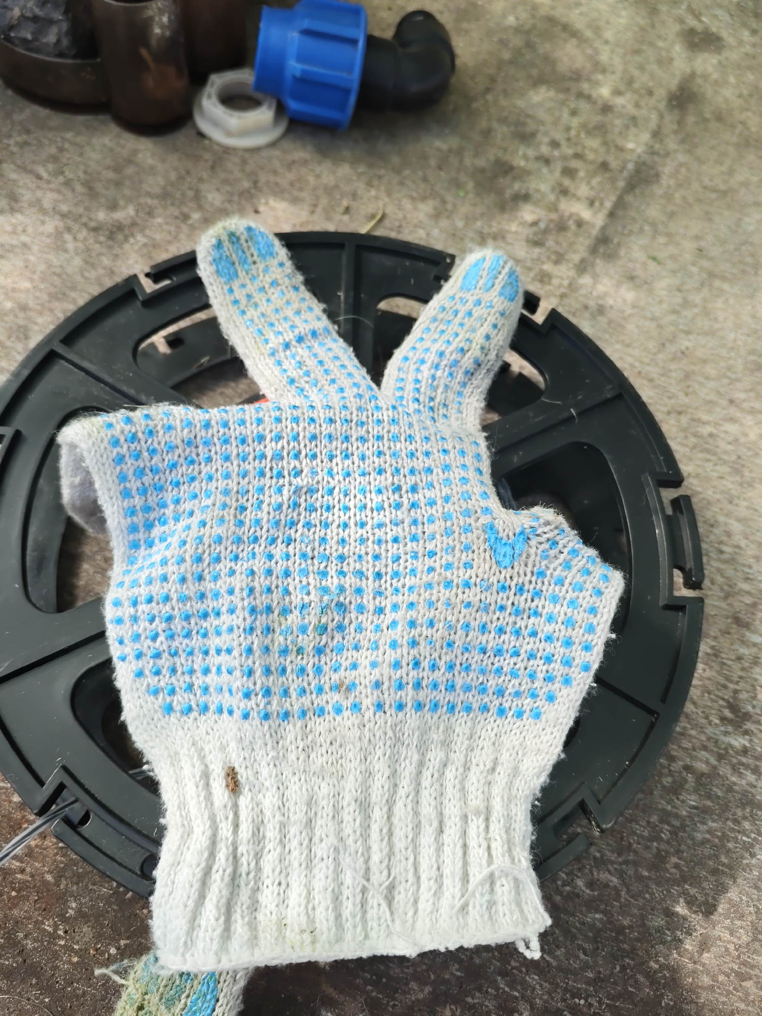 A close-up shot shows a white knit glove with blue dots, lying on a black circular object with a ridged edge. The glove appears to be a work glove, with its fingers spread open and pointing upwards. In the blurred background, various objects are visible, including a blue plastic fitting, dark metallic cylinders, and a white plastic nut. The ground beneath the objects is a rough, grey surface, possibly concrete or asphalt, with some dirt and debris. The lighting suggests an outdoor setting, possibly in the city of Rostov-on-Don, Russia.