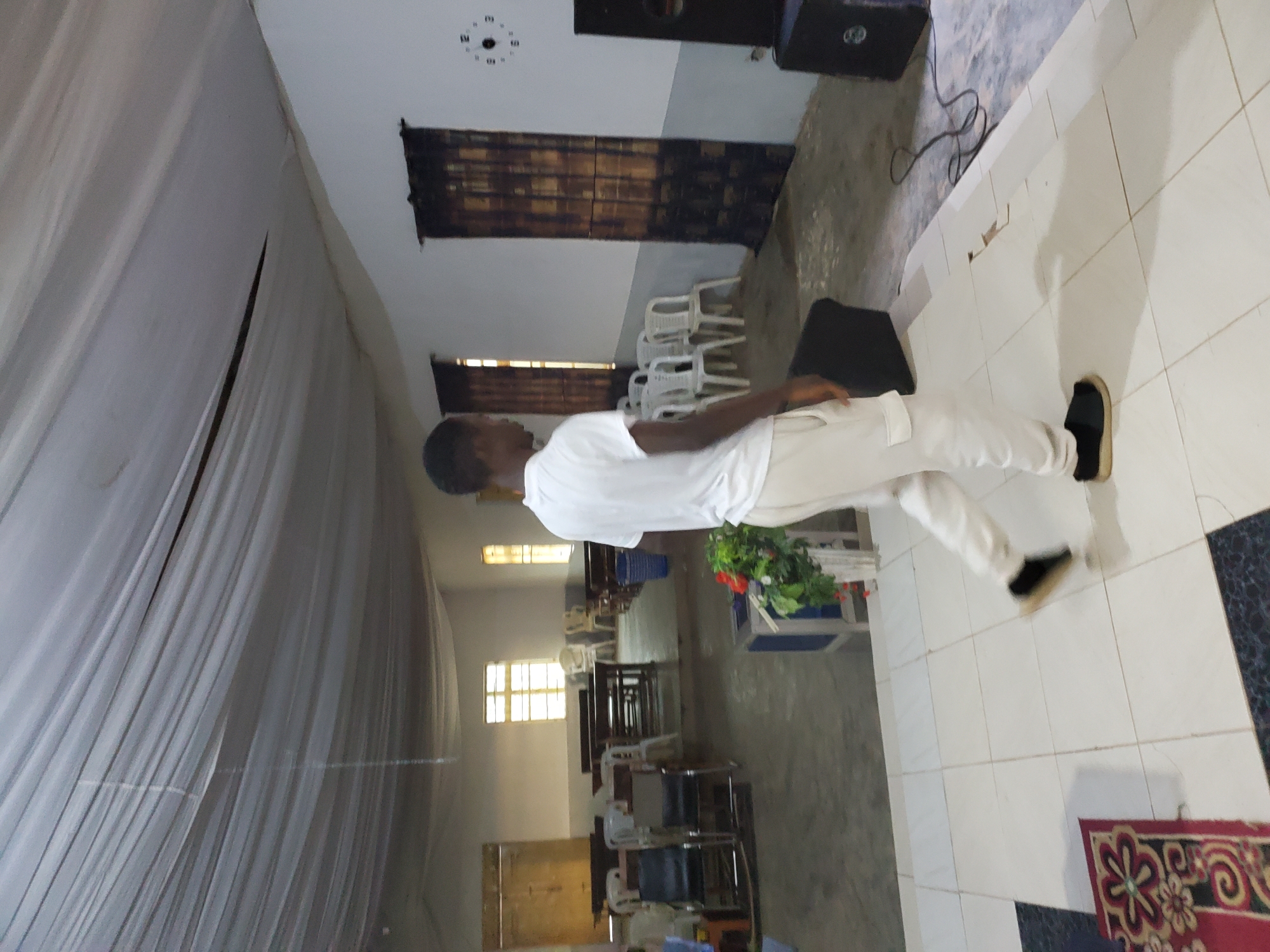 The image depicts an indoor scene, likely a hall or meeting room, in Kuru Karama, Nigeria. The room has a high ceiling draped with white fabric, creating a textured canopy. White plastic chairs are arranged in rows, and several wooden tables are also visible, suggesting a classroom or event setup. Natural light enters through windows with dark curtains. A young Black male, wearing a white t-shirt and light-colored pants, is walking across a tiled floor towards the right side of the frame. He is in motion, with his legs blurred. A black speaker and a clock on the wall are also present. A decorative vase with red and green flowers sits on a platform to the left of the moving figure.