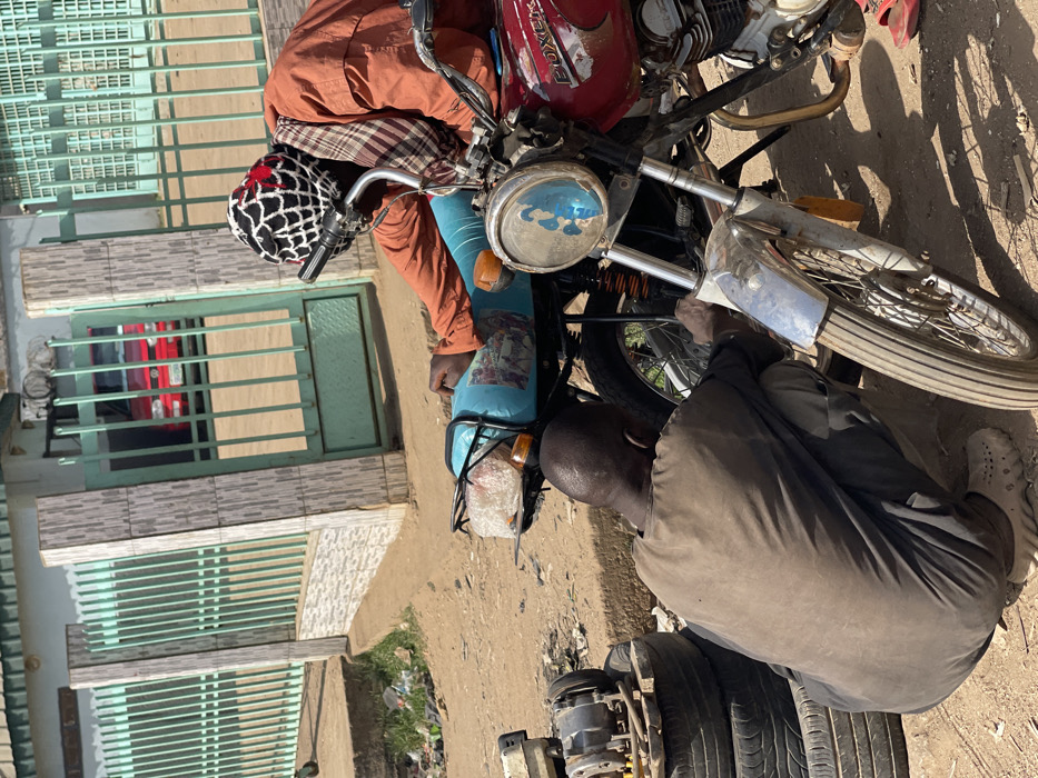 In Chikaji, Nigeria, an outdoor scene depicts two adult males engaged in motorcycle repair on a dirt ground. The individual on the left, wearing a dark brown tunic and trousers with light-colored clogs, is crouched, facing away from the camera, attending to the rear wheel area of a light blue motorcycle. Adjacent to him on the left, a stack of three tires supports a metallic engine component. The second individual, positioned on the right, wears an orange long-sleeved shirt, a plaid neck wrap, and a black and white mesh-patterned beanie. He leans over a red "Boxer" brand motorcycle, its front headlight adorned with a blue sticker, focusing on its front section. Red sandals are visible near his feet. The background features a light green metal fence and gate with tiled pillars. Through the open gate, a red vehicle is visible. Scattered refuse is present on the dirt surface to the far left. The scene is brightly lit by natural sunlight, casting shadows.