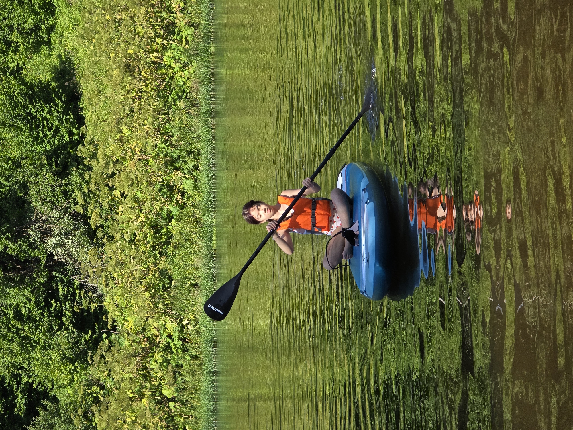 A young girl sits on a blue paddleboard in the middle of a calm, green body of water, holding a paddle. She is wearing an orange life vest and appears to be paddling. The paddleboard is in focus, as is the girl. In the background, a lush green forest lines the edge of the water. The scene is brightly lit by the sun, casting reflections on the water's surface. The location is the Voinskaya Chast' Mchs, Russia.
