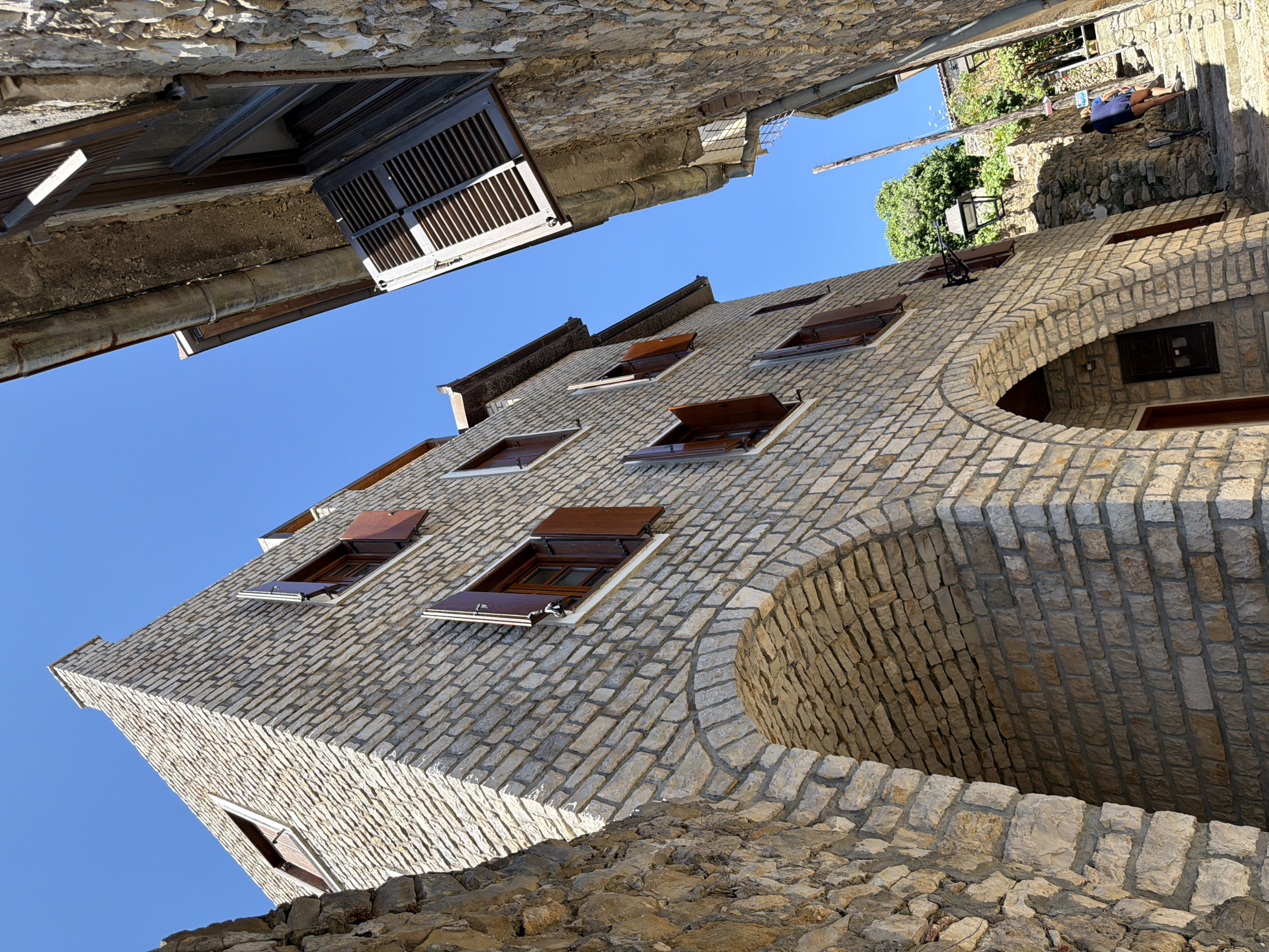 A low-angle shot captures a narrow street in the old town of Ulcinj, Montenegro, with two stone buildings flanking the pathway. The building on the left, constructed of light-colored stone bricks, features arched entrances at its base and several windows with dark wooden shutters, some of which are open. The building on the right, also made of stone, is positioned at an angle, with a portion of its facade and a sloped roof visible. A person in a blue shirt and dark shorts is walking up a stone staircase that ascends between the two buildings, with a bicycle leaning against a wall nearby. Lush green trees and more stone structures are visible in the background under a clear blue sky.