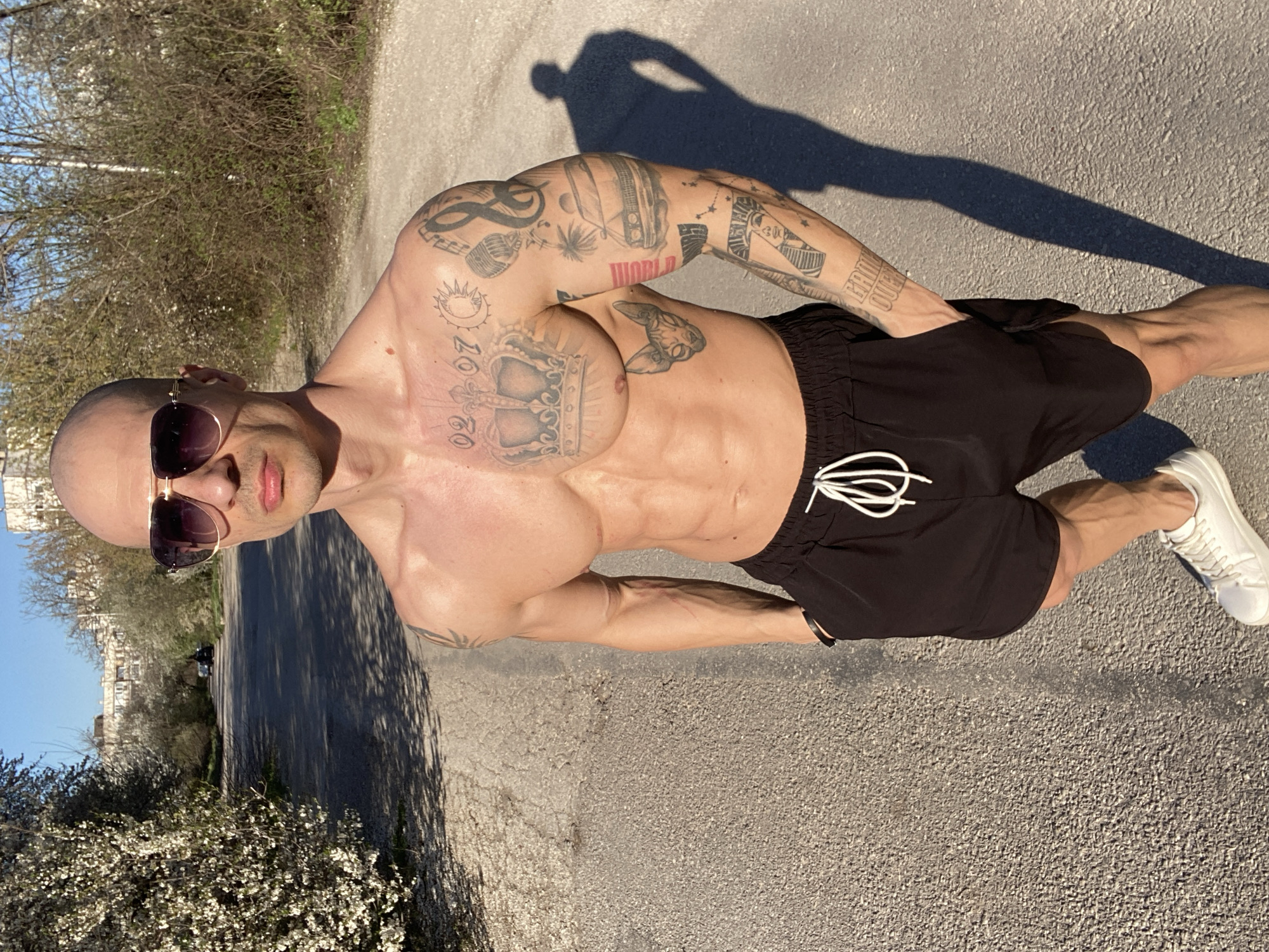 A bald man wearing sunglasses and black shorts stands on an asphalt path. He has numerous tattoos on his upper body. The path is lined with trees and flowering bushes. His shadow is cast on the path behind him. Buildings are visible in the background. The image appears to be taken outdoors in Pleven, Bulgaria.